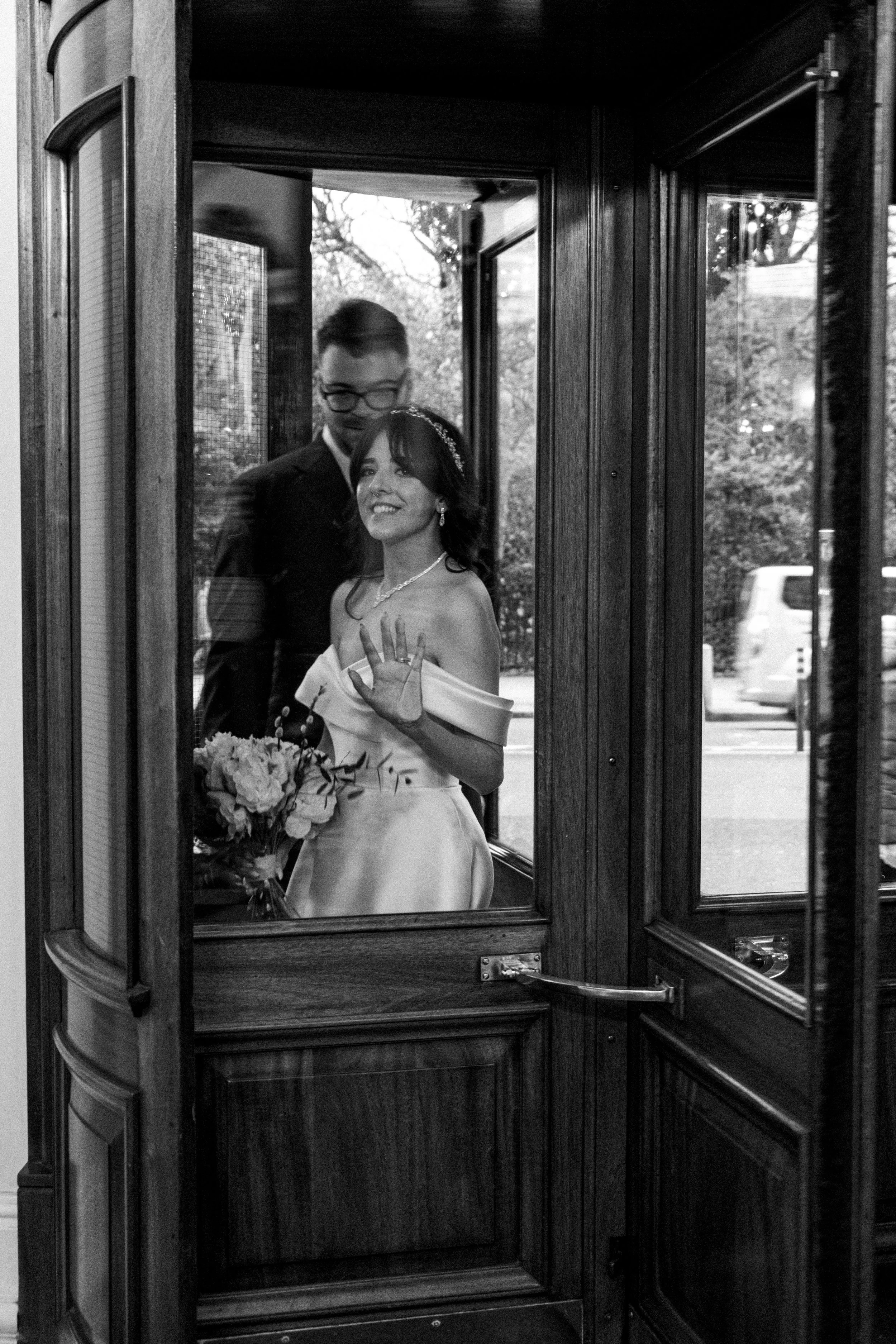 Black and white photo of a bride waving through a glass door, holding a bouquet of flowers, with a groom standing behind her.