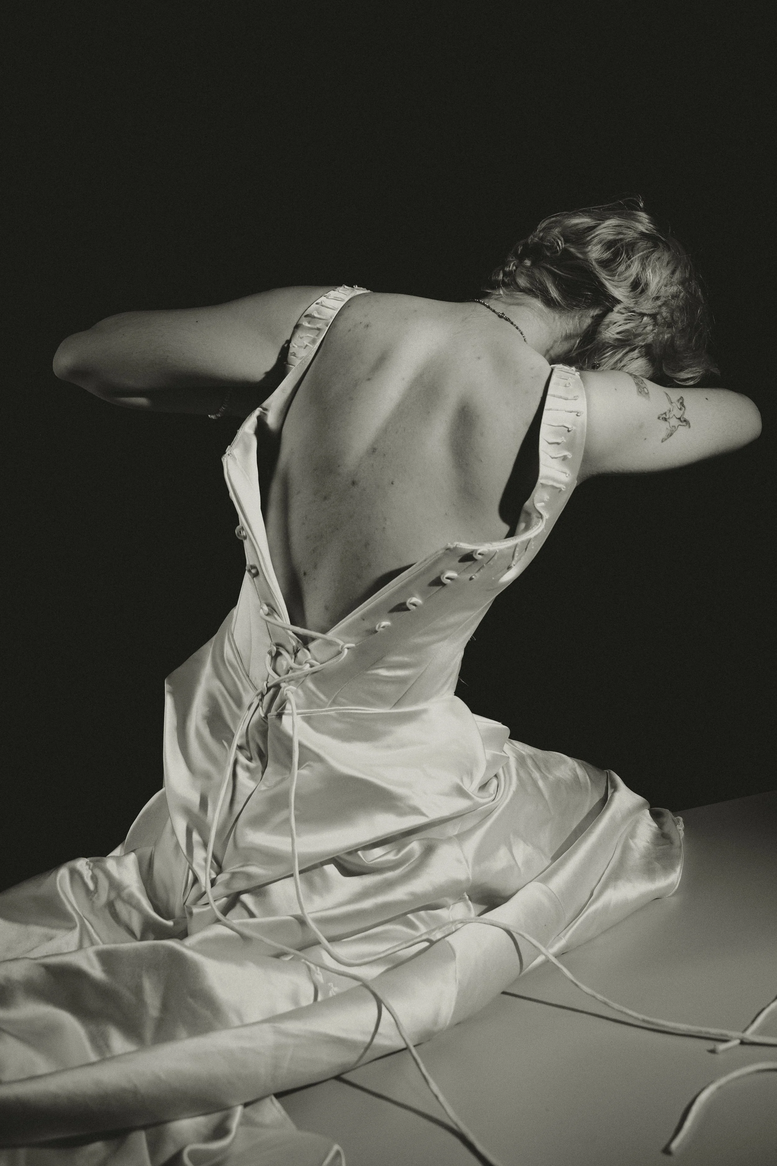 A black and white photo of a woman sitting on the floor with her back to the camera, wearing a satin dress with open back, her arms are crossed behind her head, and several cords are draped around her. She has tattoos on her arm and wavy hair.
