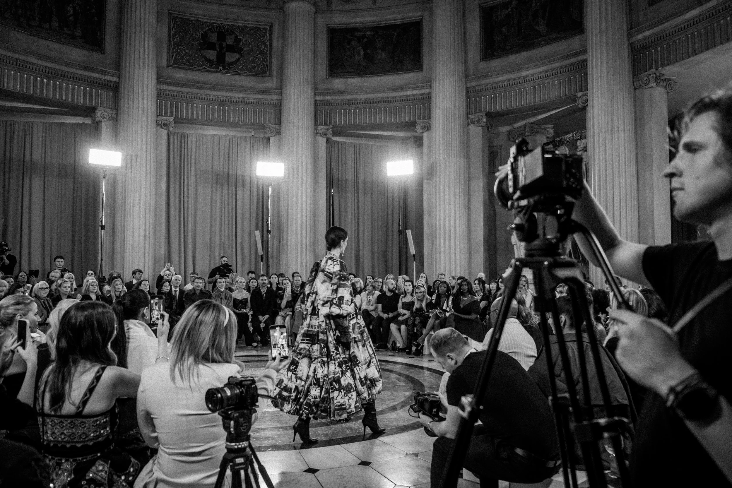 A fashion runway show with a model walking in front of an audience in an ornate hall, surrounded by photographers and spectators capturing the event.