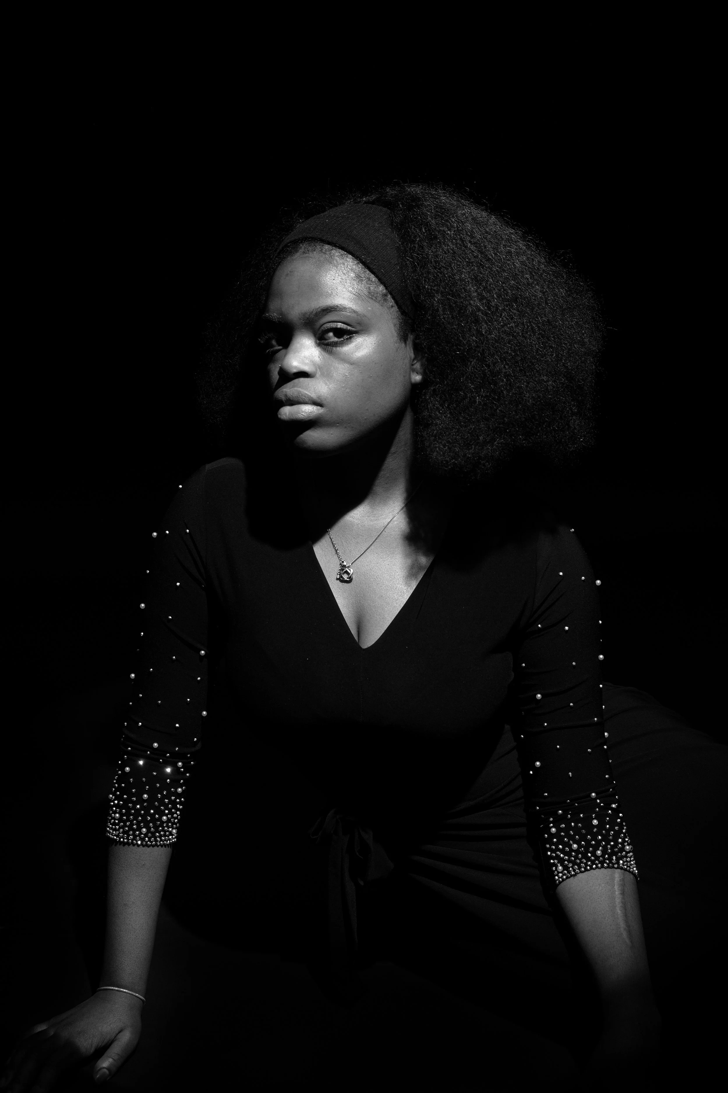 Black and white portrait of a young woman with natural hair, wearing a dark top with embellishments, sitting in a dark environment.