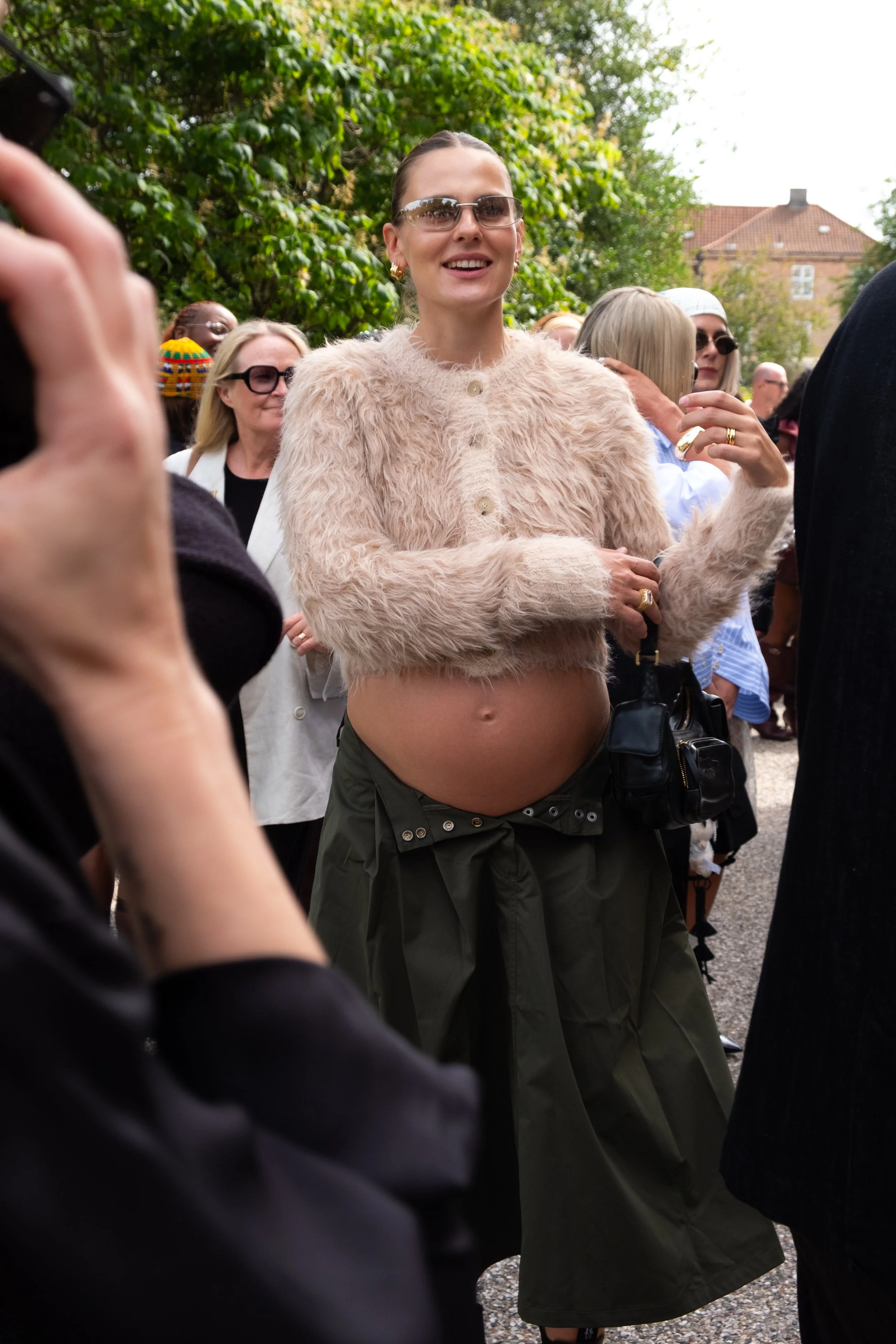 A woman wearing a fuzzy pink jacket, sunglasses, and green pants stands among a crowd outdoors.