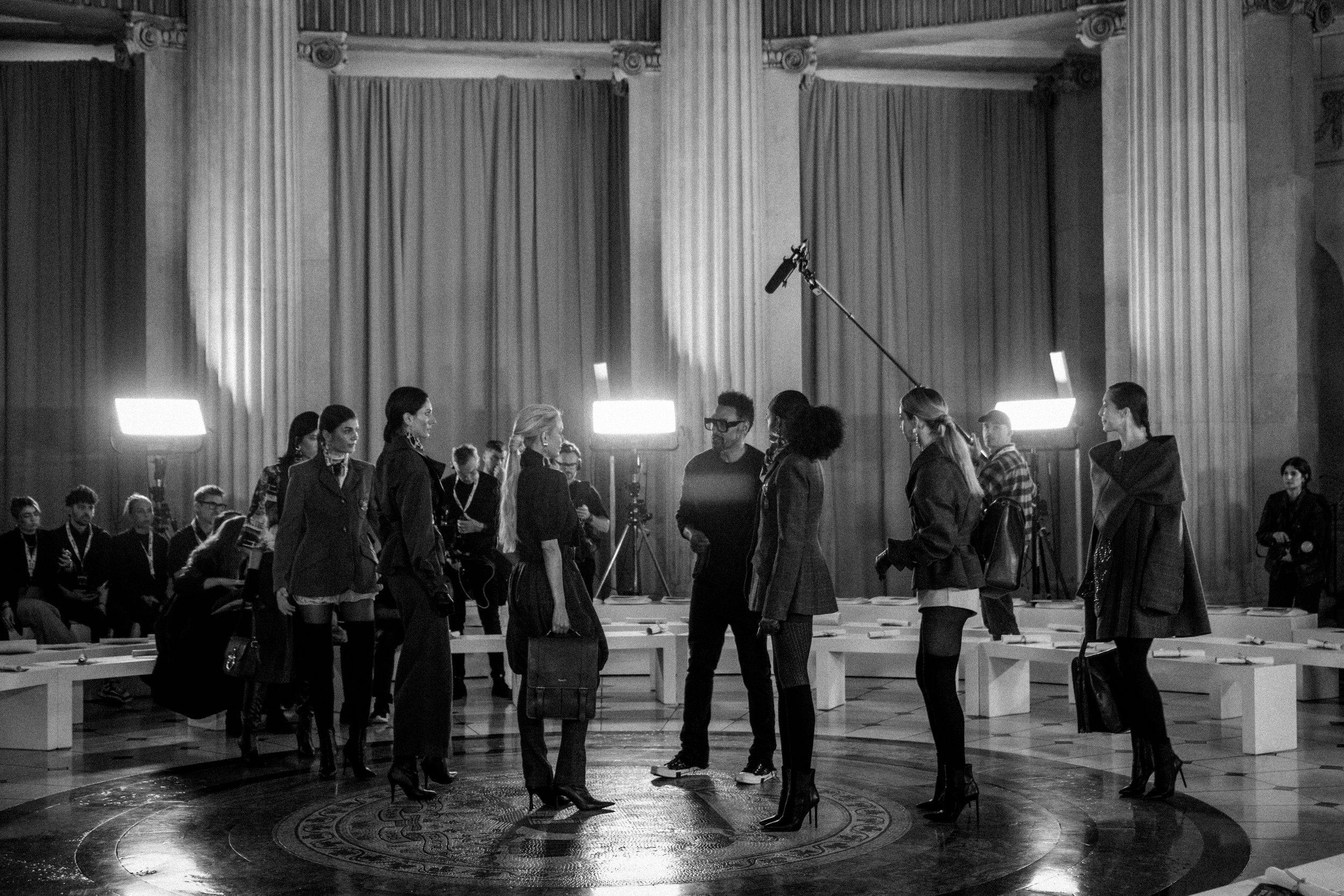 A black and white photograph of a film or television production set inside a large, classical building with tall columns and draped curtains. Several women in stylish attire stand in a circle around a man who appears to be giving instructions, while 