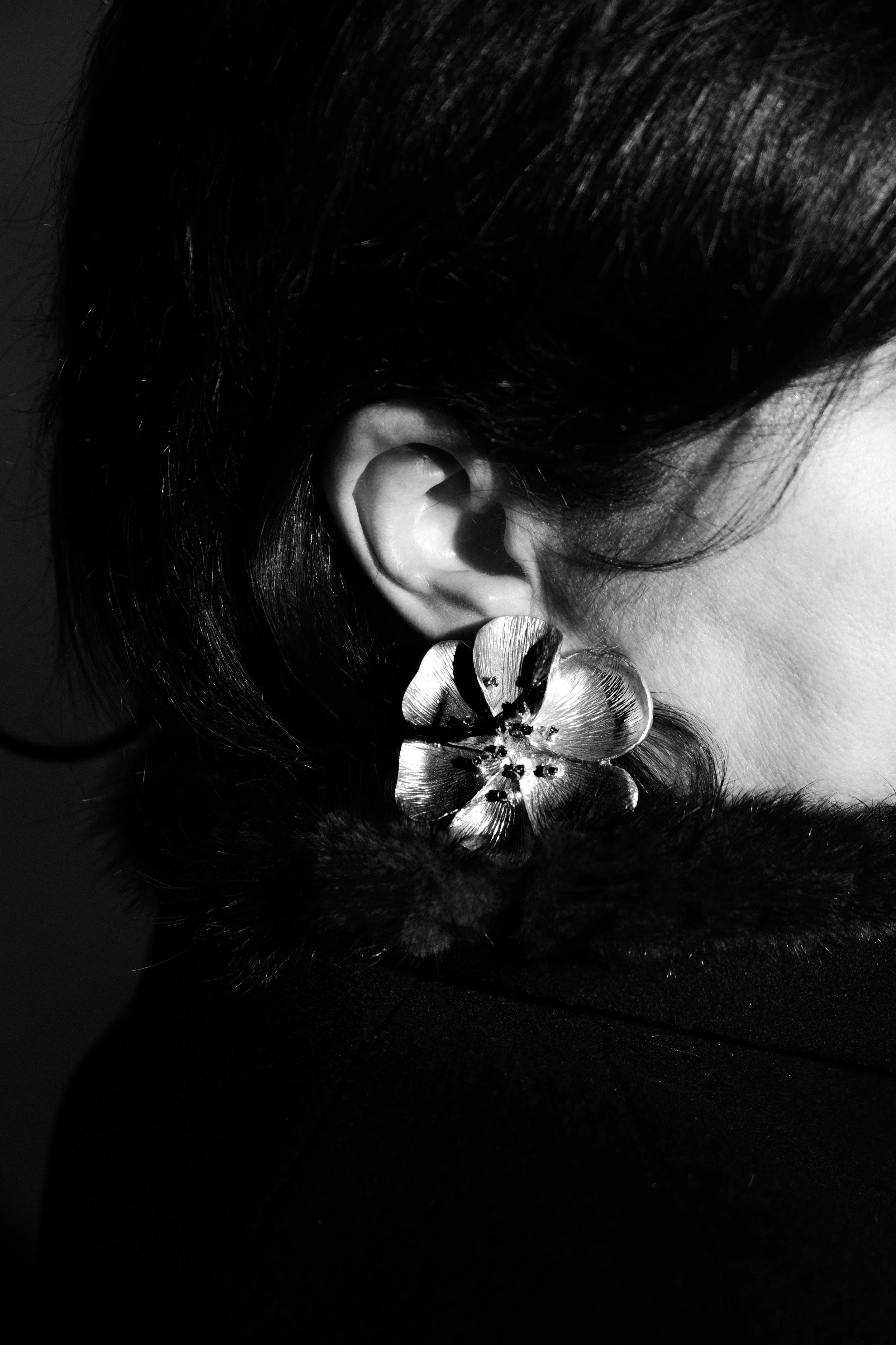 A black and white close-up of a woman's ear with a large flower earring and her dark hair, wearing a black fur collar.