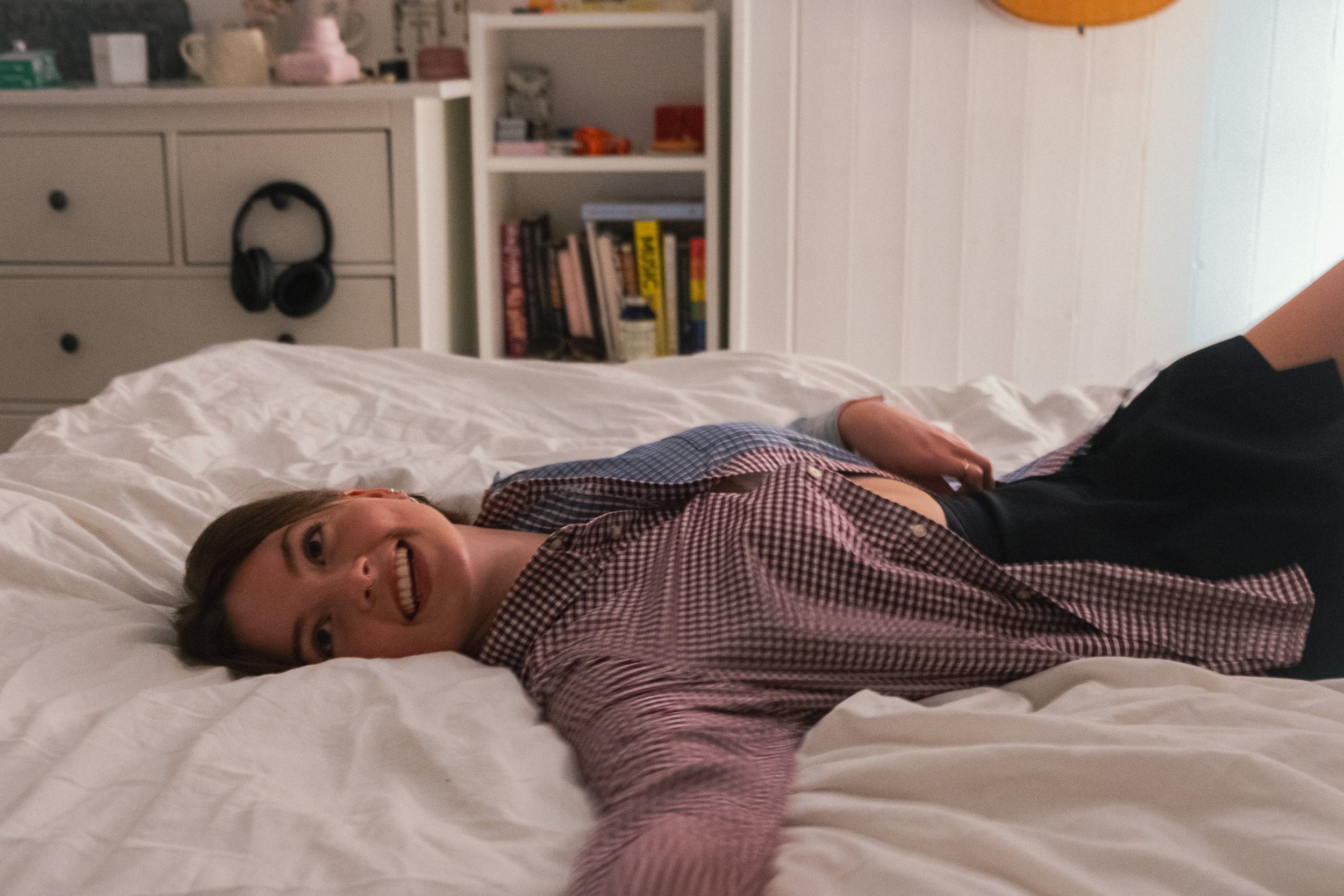 A woman lying on her side on a bed, smiling at the camera, in a bedroom with white walls, a white dresser with black knobs, a bookshelf with books, and a pair of black headphones hanging on the dresser.