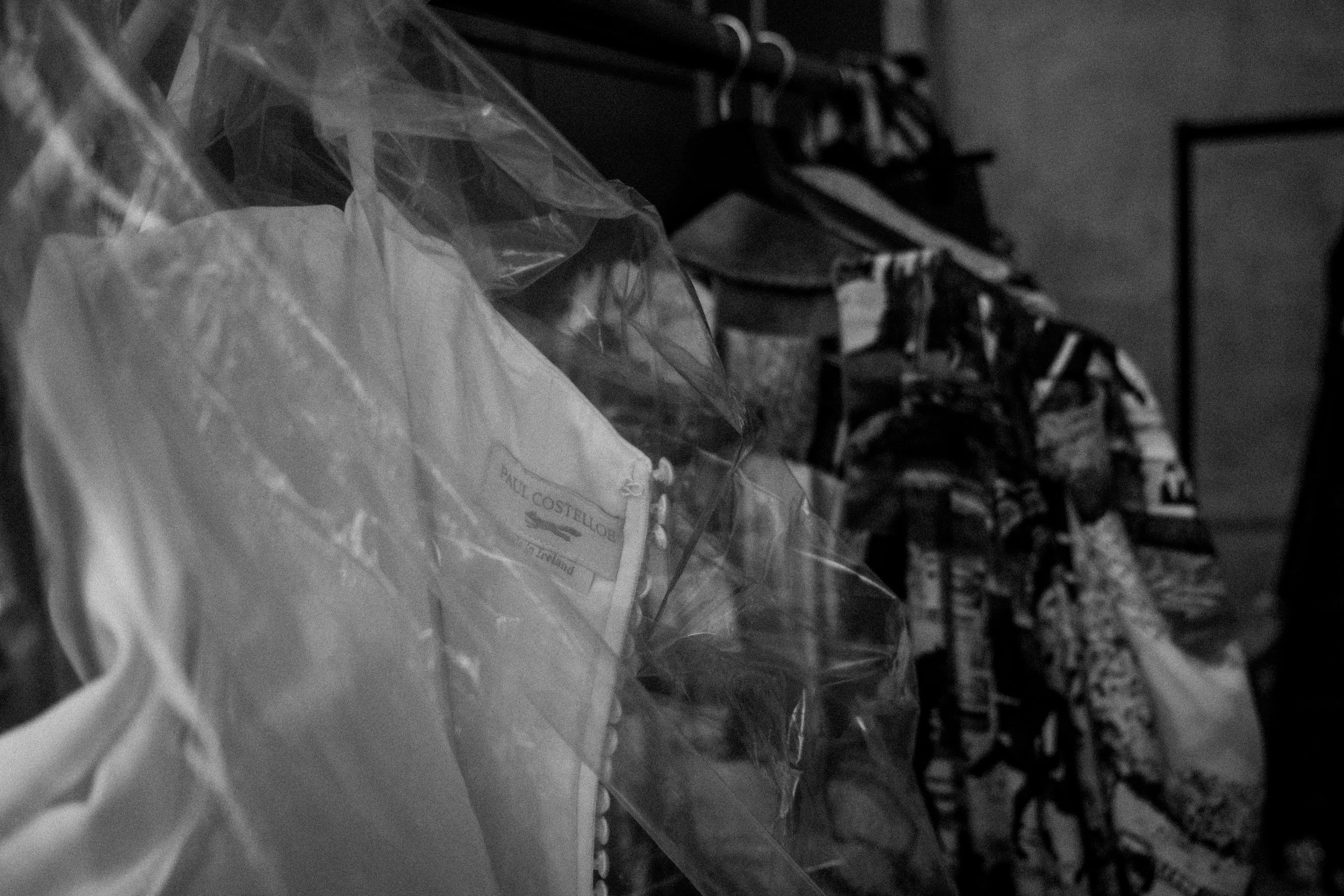 Hanging clothes in a closet, with a focus on a packaged garment labeled 'Paul Costelloe.' The clothes include various patterned fabrics.