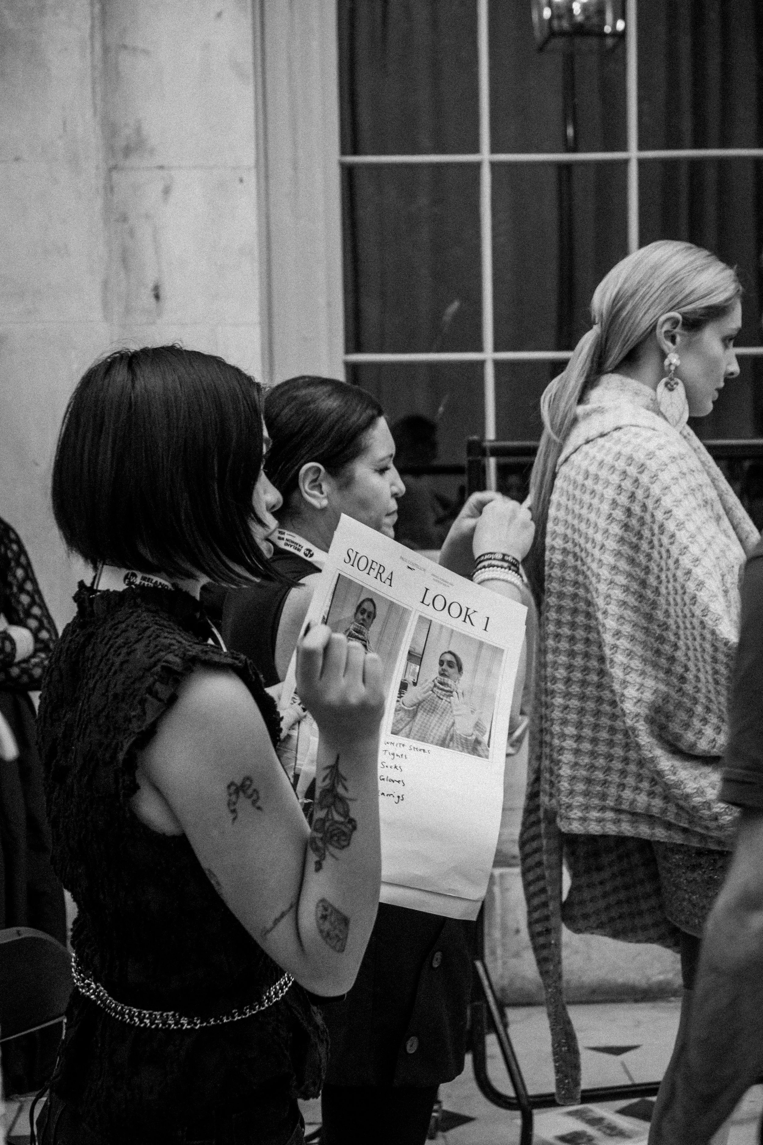A woman with tattoos and a black lace top holding a clipboard with a photo and notes, standing in line at an indoor event.