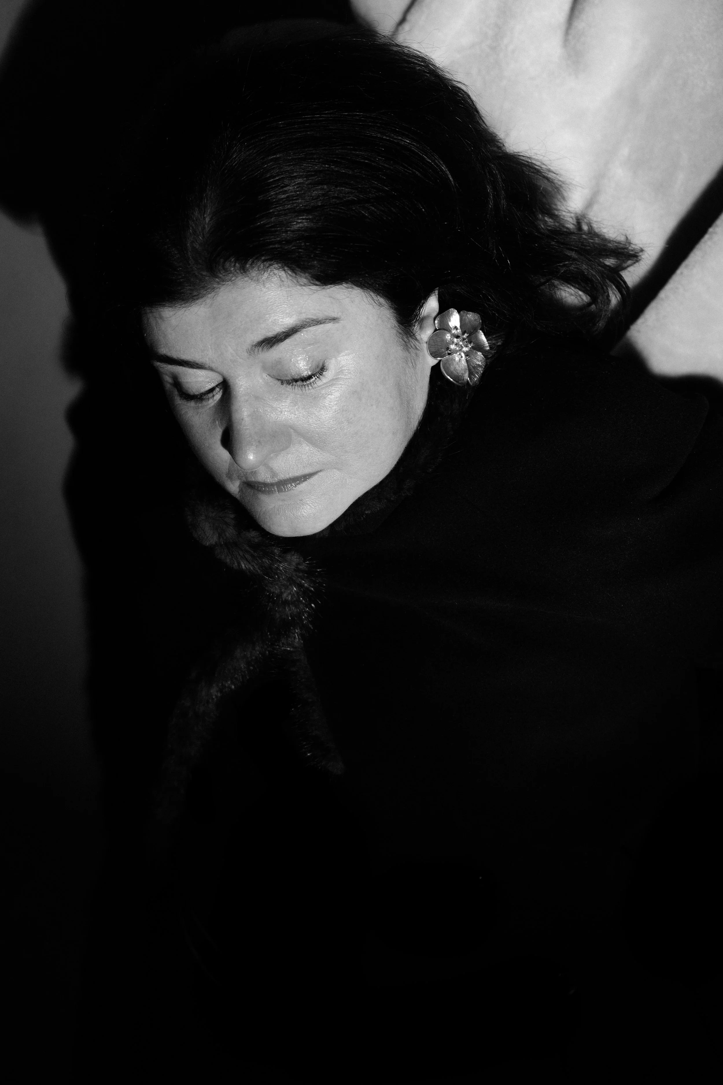 A black and white portrait of a woman with dark hair, closed eyes, and floral earrings, lying down with a contemplative expression.