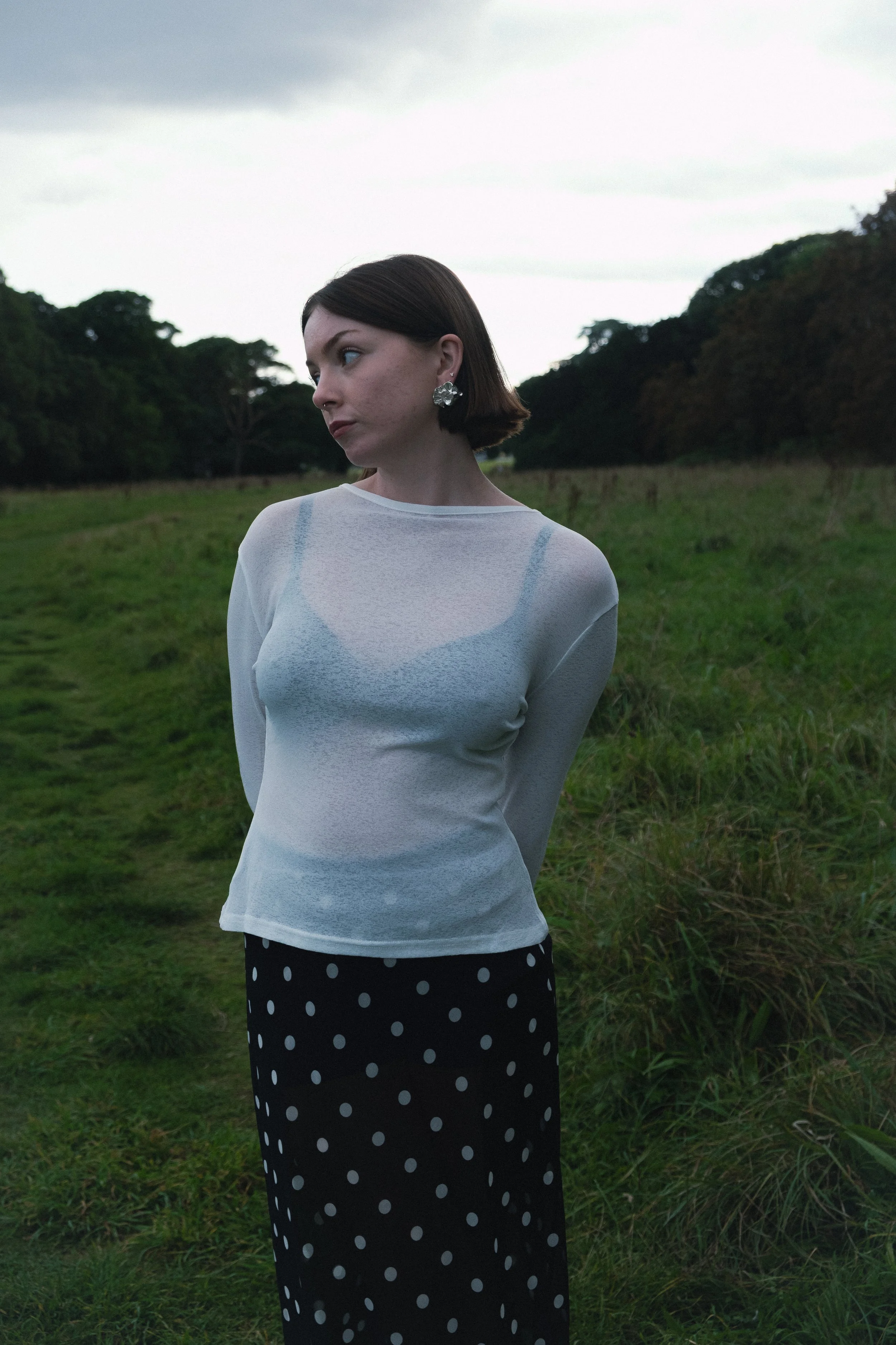 A woman with short brown hair and earrings stands in a grassy field with trees in the background, under a cloudy sky, wearing a sheer white top and a black polka dot skirt.