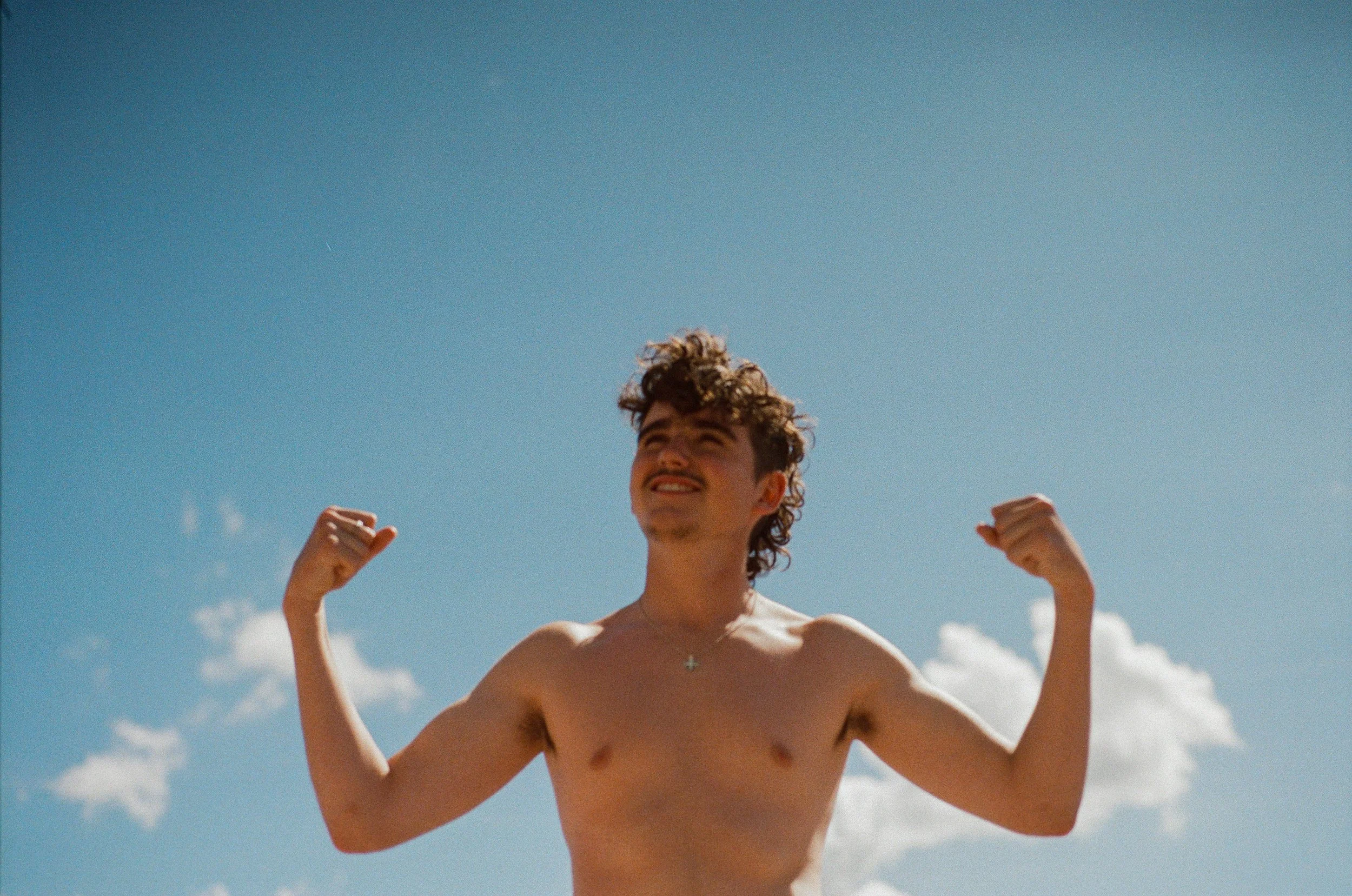 A shirtless young man with curly hair smiling and flexing his biceps outdoors under a blue sky with some clouds.