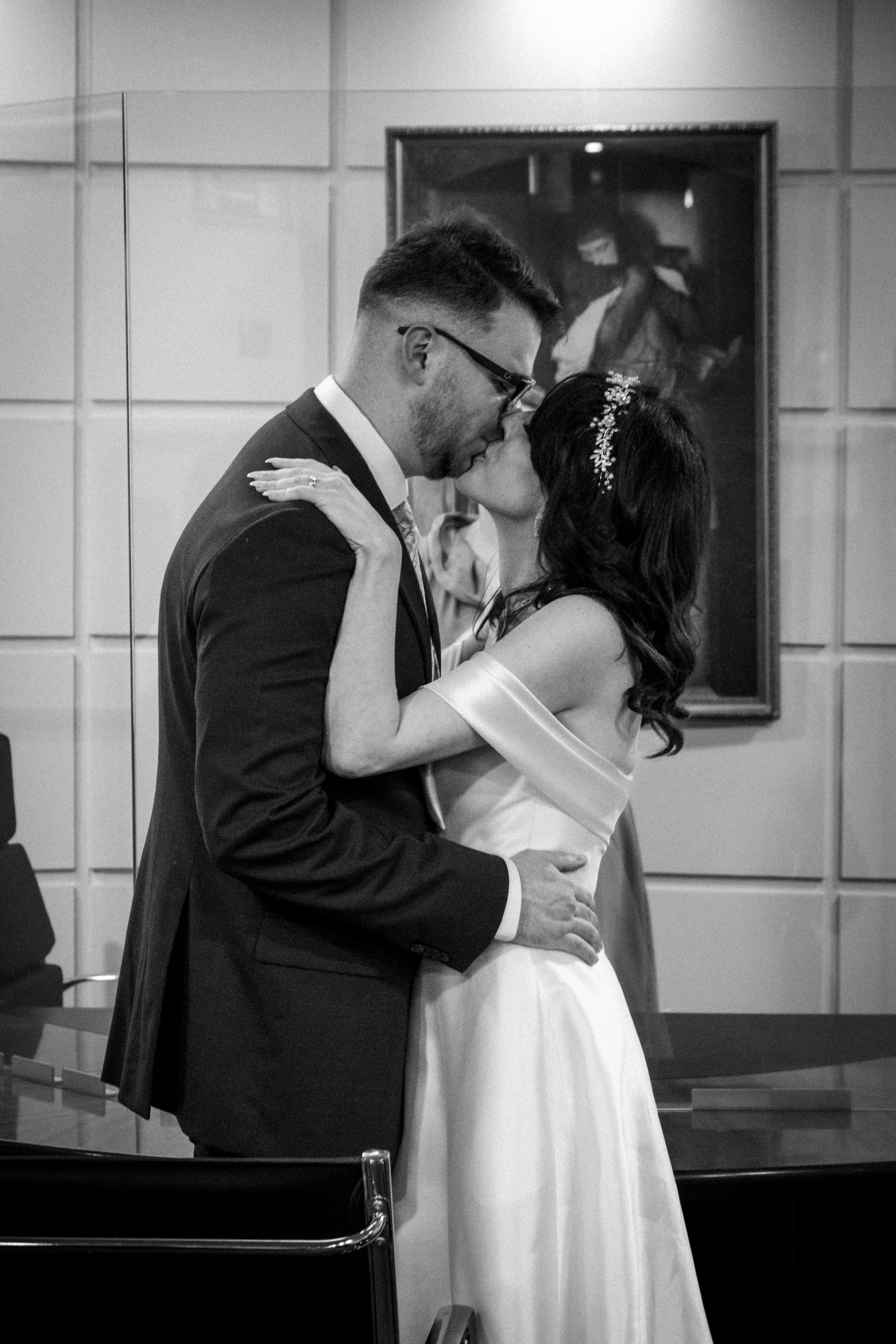 A couple in formal wear kissing during a wedding ceremony, with a painting behind them on the wall.