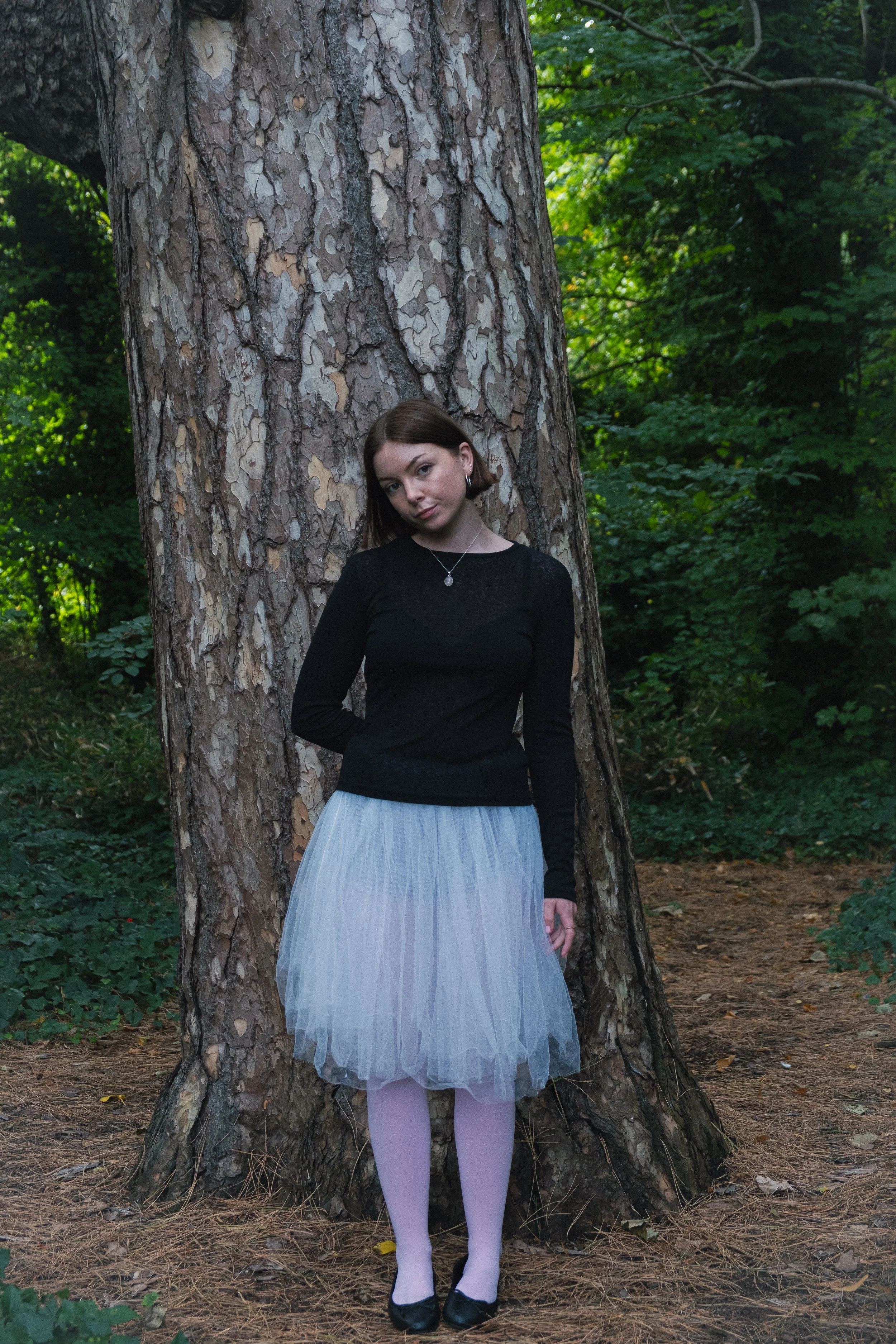 A young woman in a black long-sleeve top, white tulle skirt, and pink tights stands leaning against a large tree in a wooded area during daytime.