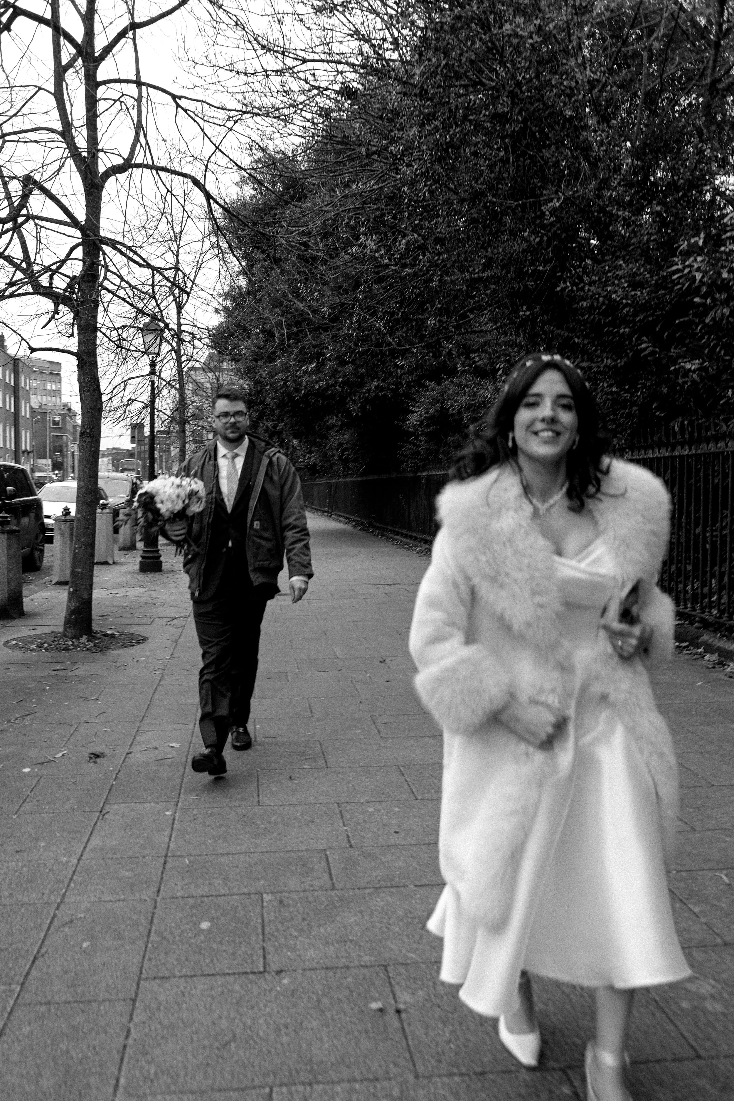 A black-and-white photo of a woman in a wedding dress and fur coat walking on a sidewalk, smiling at the camera, with a man in a suit and tie holding a bouquet of flowers walking behind her.