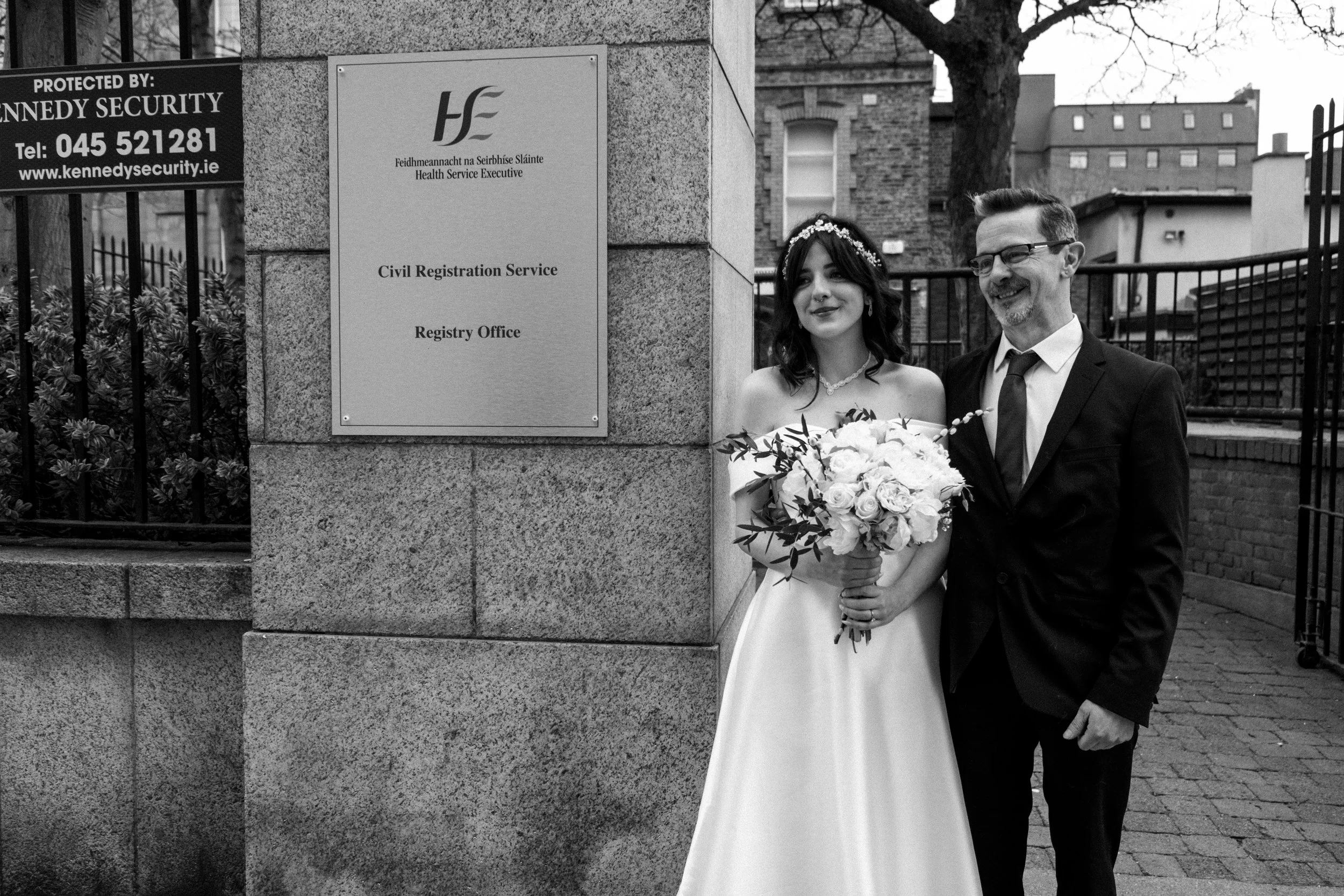A bride and groom pose for a photo outside the Civil Registration Service office, with the bride holding a bouquet of flowers and wearing a wedding dress, and the groom in a suit and tie. There is a sign on the stone wall with Irish and English text.