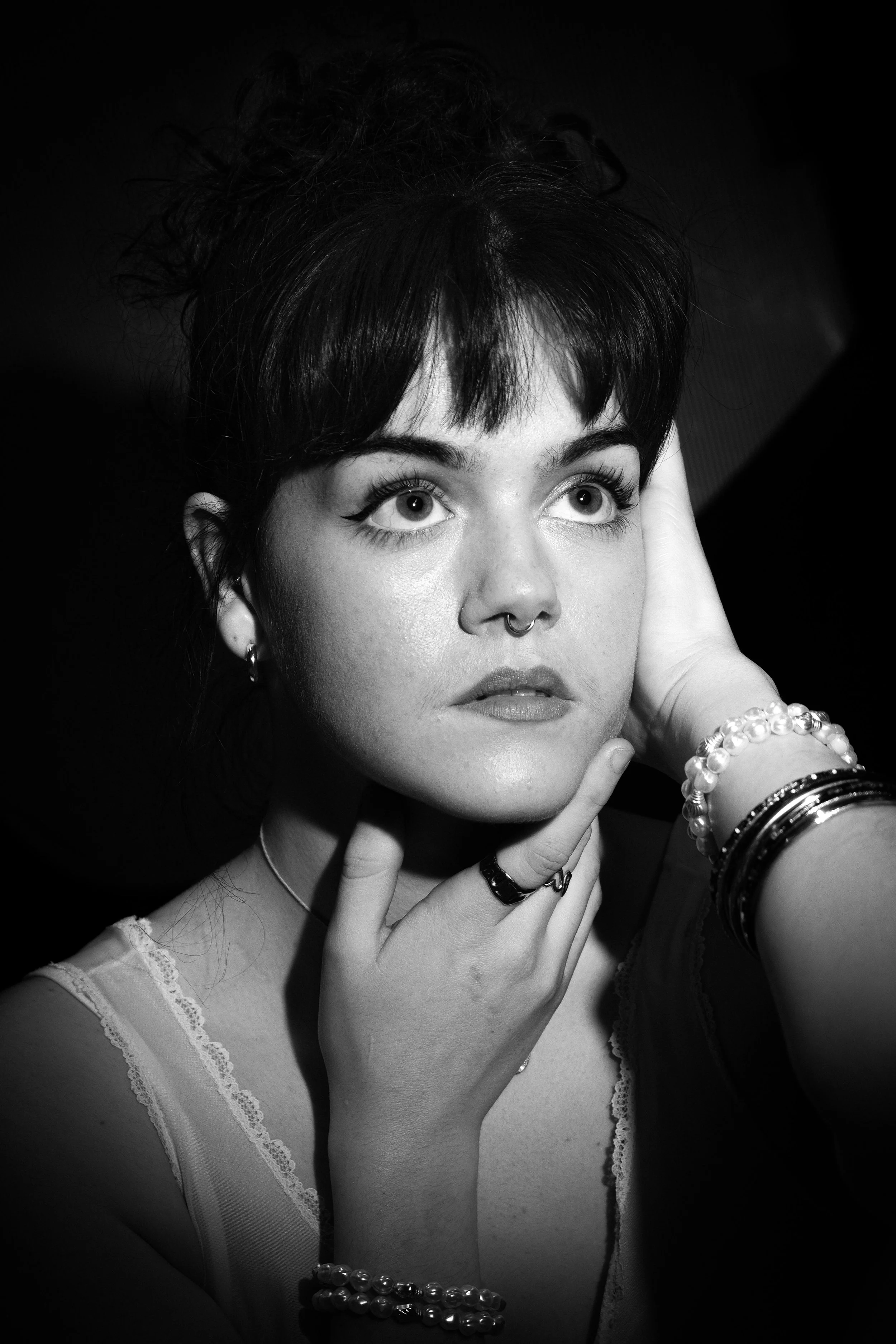 Black and white portrait of a young woman with short dark hair, looking upwards with wide eyes, touching her face with her left hand.