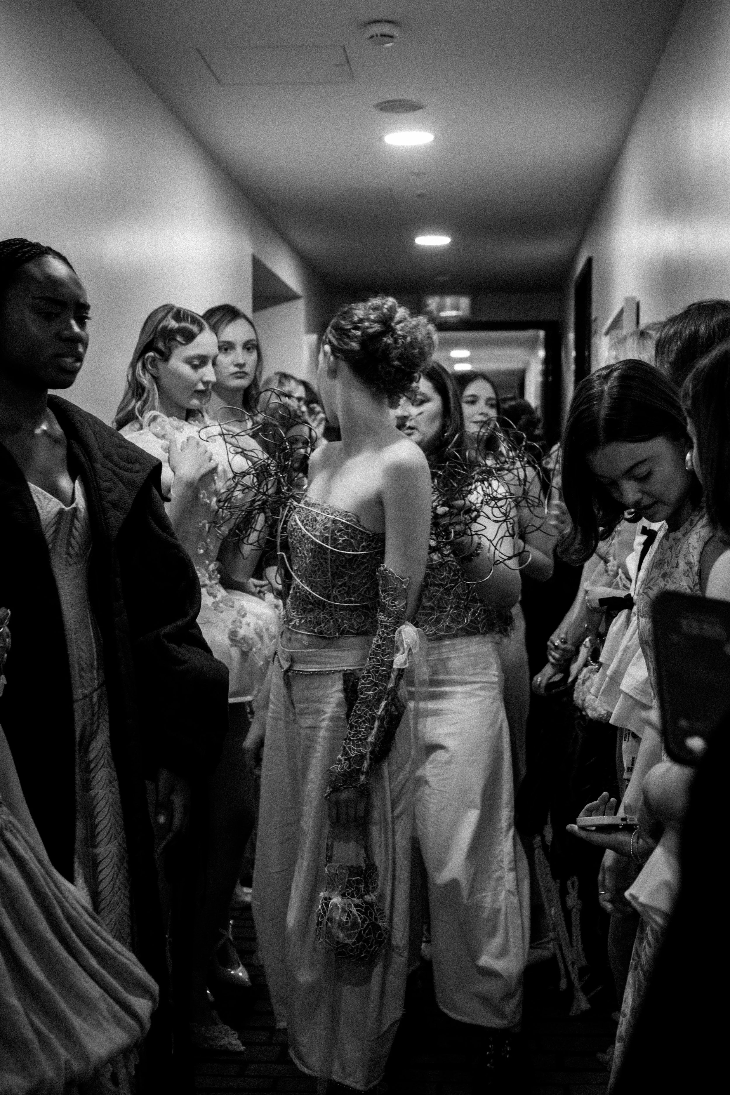 Group of women in a hallway wearing artistic, elaborate clothing, with one woman in the center dressed in detailed, textured attire and others looking on.