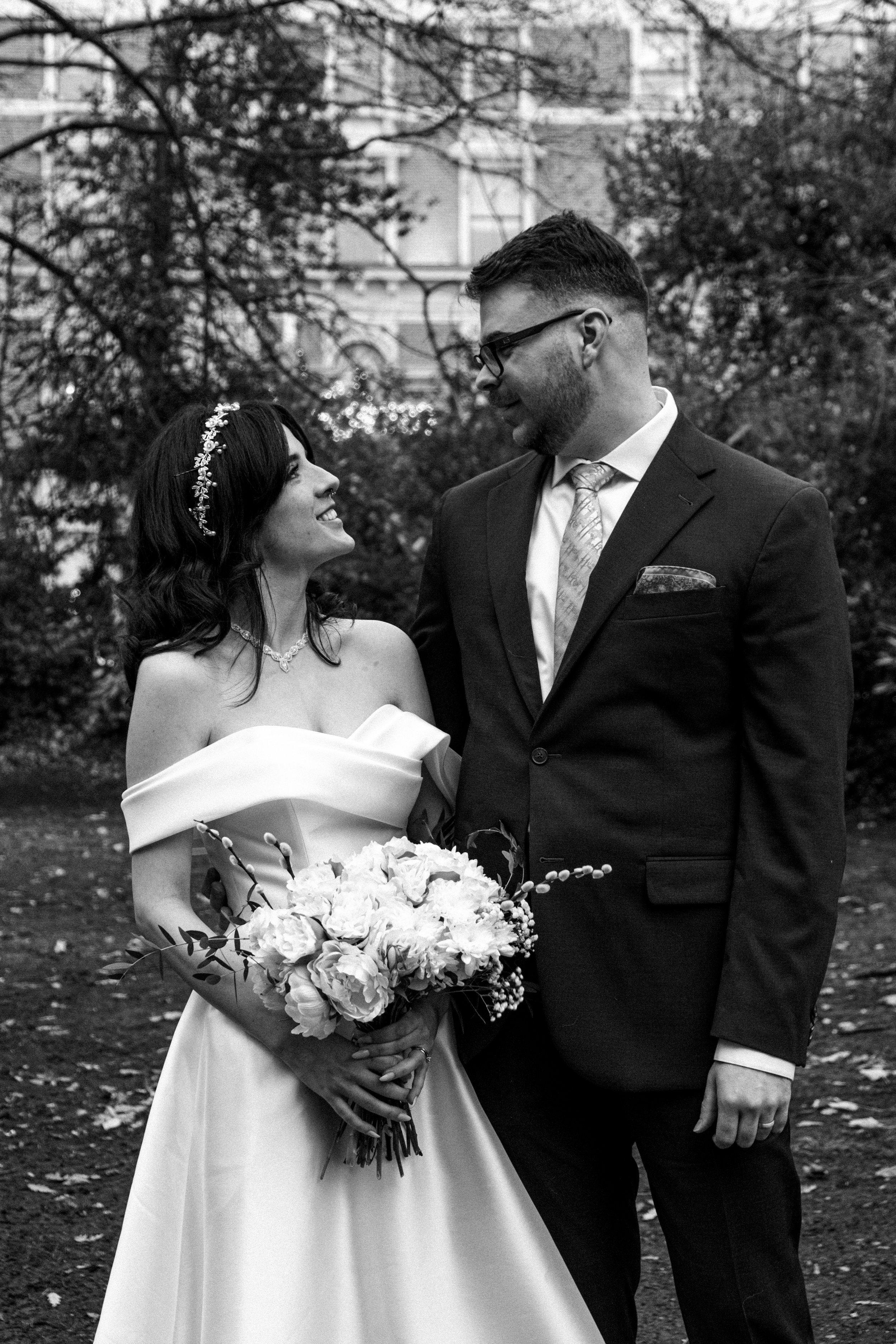 A bride and groom smiling at each other outdoors with trees and buildings in the background, black and white photo.