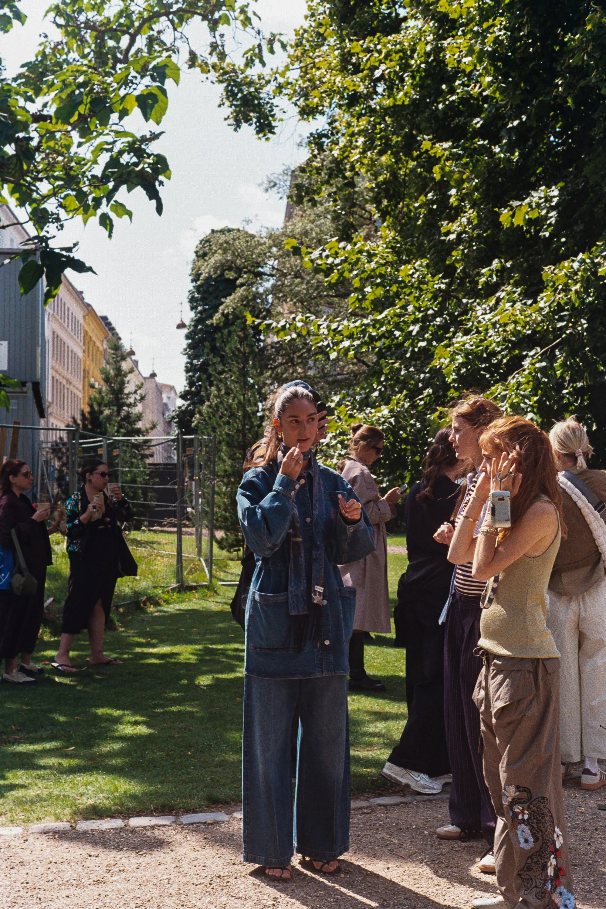 Group of women, some on cell phones, gathered outdoors on a grassy area with trees, in a city setting on a sunny day.
