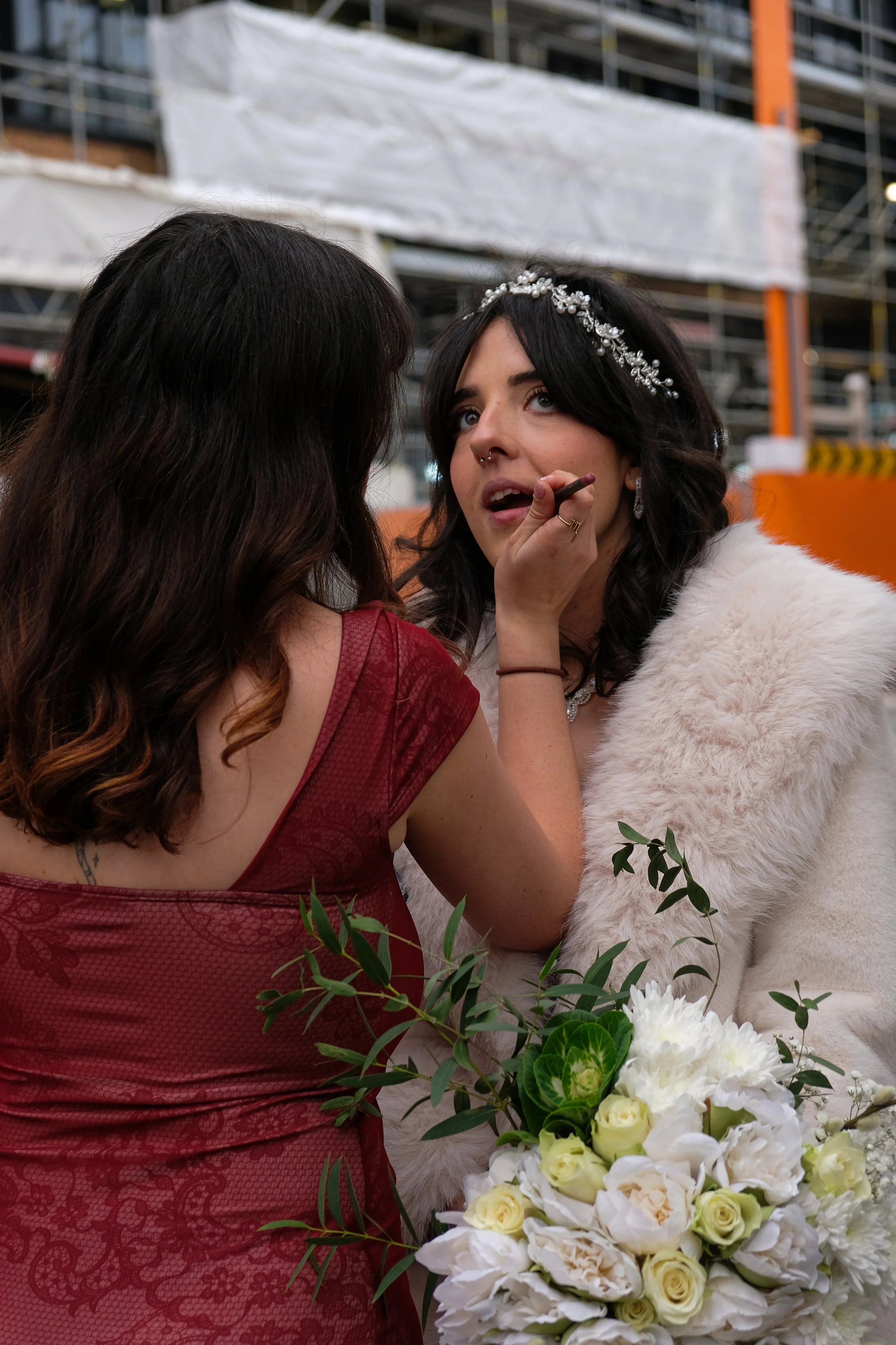 A bride with dark hair and a floral headband receives makeup application from a makeup artist at her wedding venue, with a bouquet of white roses and greenery in the foreground.