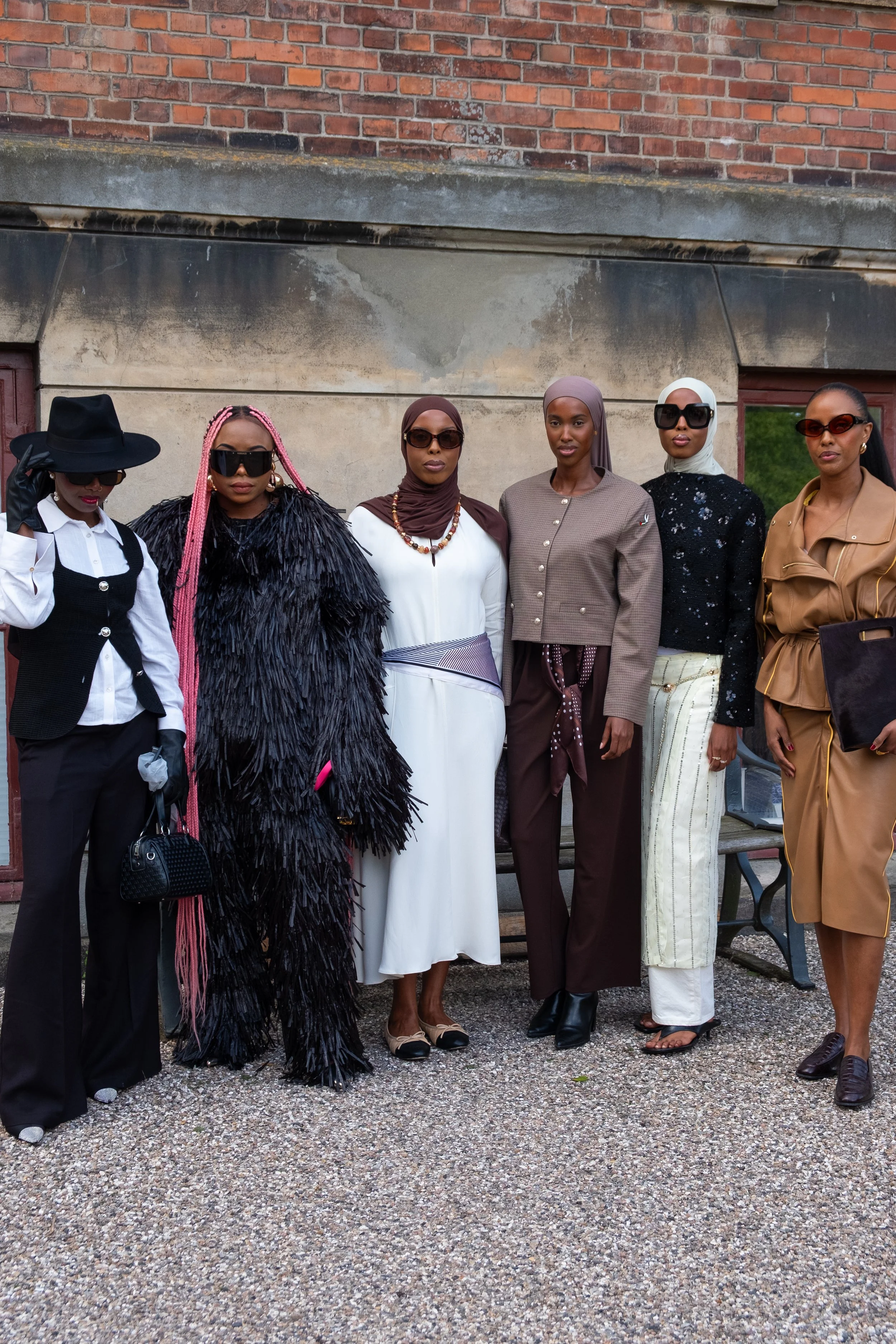 Group of seven women standing outdoors in fashionable clothing, posing for a photo in front of a brick wall.