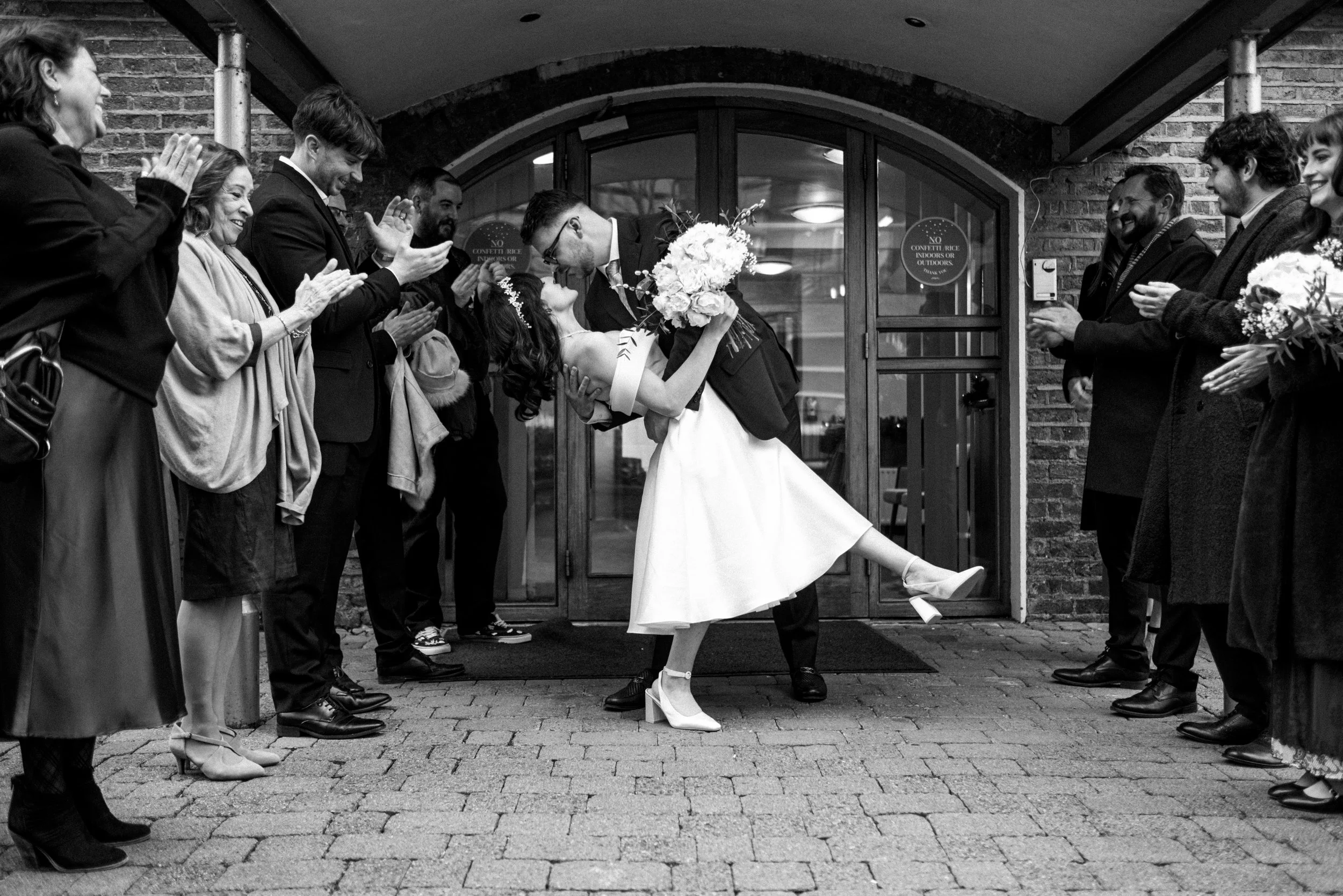 A bride and groom sharing a kiss outside a building with friends and family on either side, clapping and celebrating.
