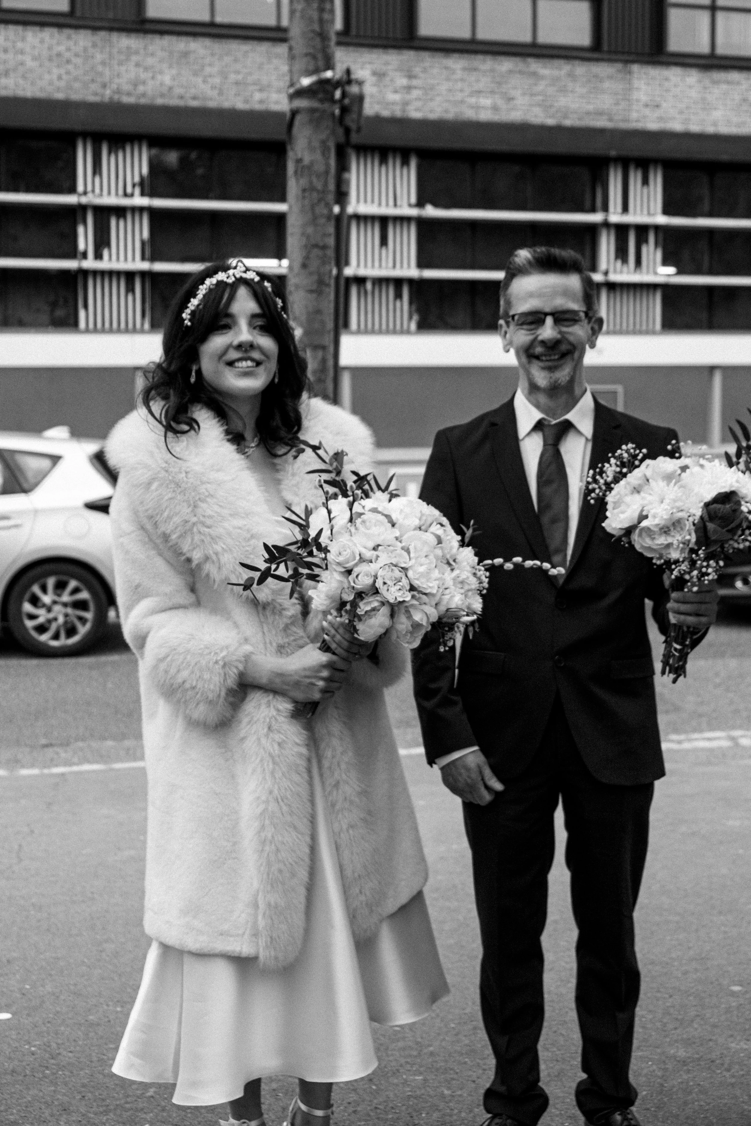 Black and white photo of a woman in a wedding dress and coat holding a bouquet standing next to a man in a suit and tie holding a bouquet, outdoors in front of a modern building with parked cars.