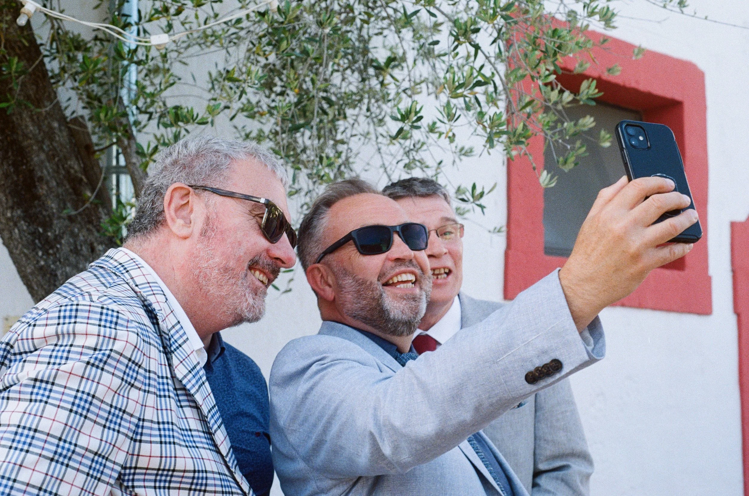Three middle-aged men with sunglasses taking a selfie outdoors near a tree and a white wall with a red window frame.