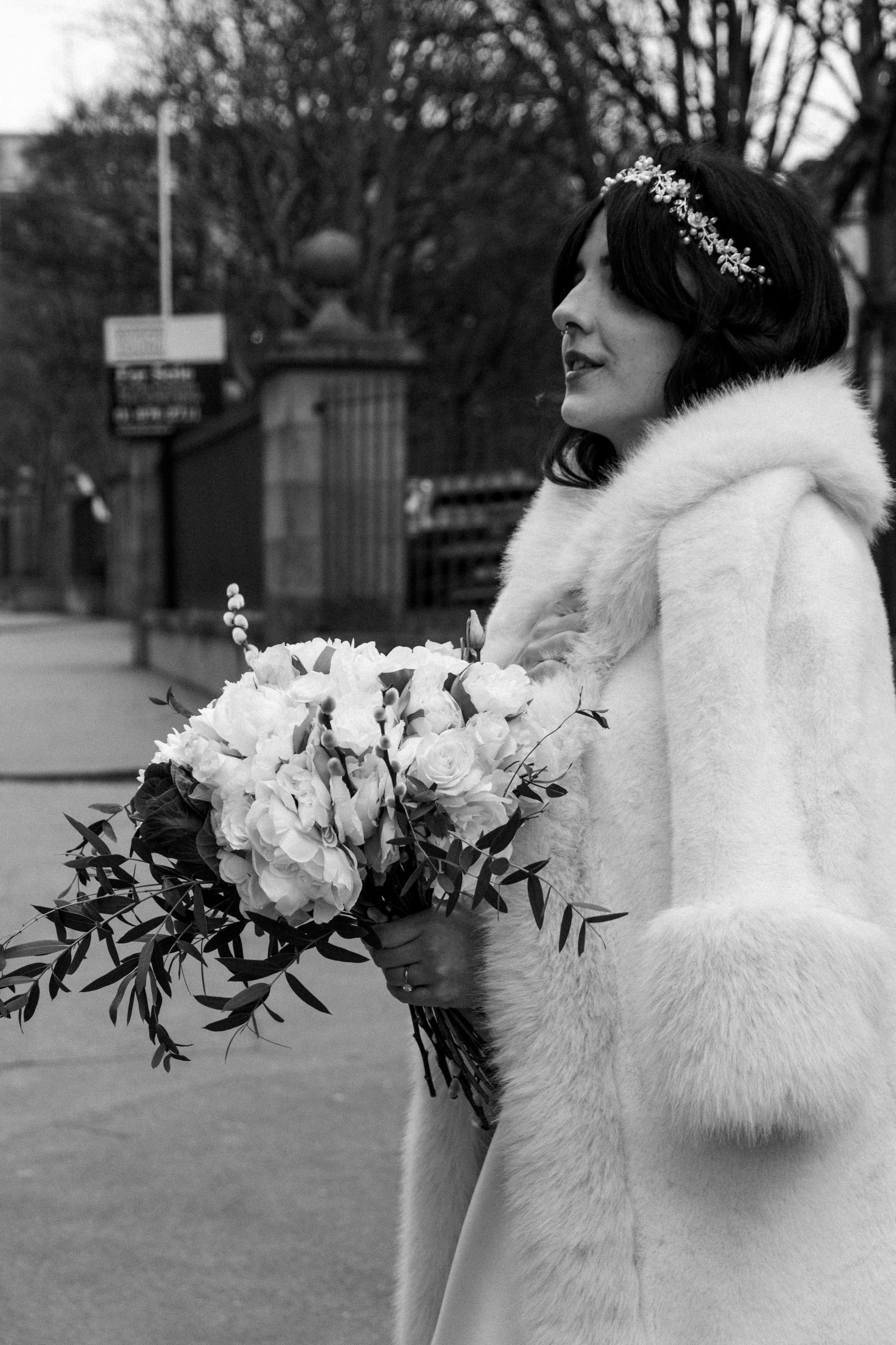 A woman in a white fur coat with a floral headband holding a bouquet of flowers in an outdoor setting.