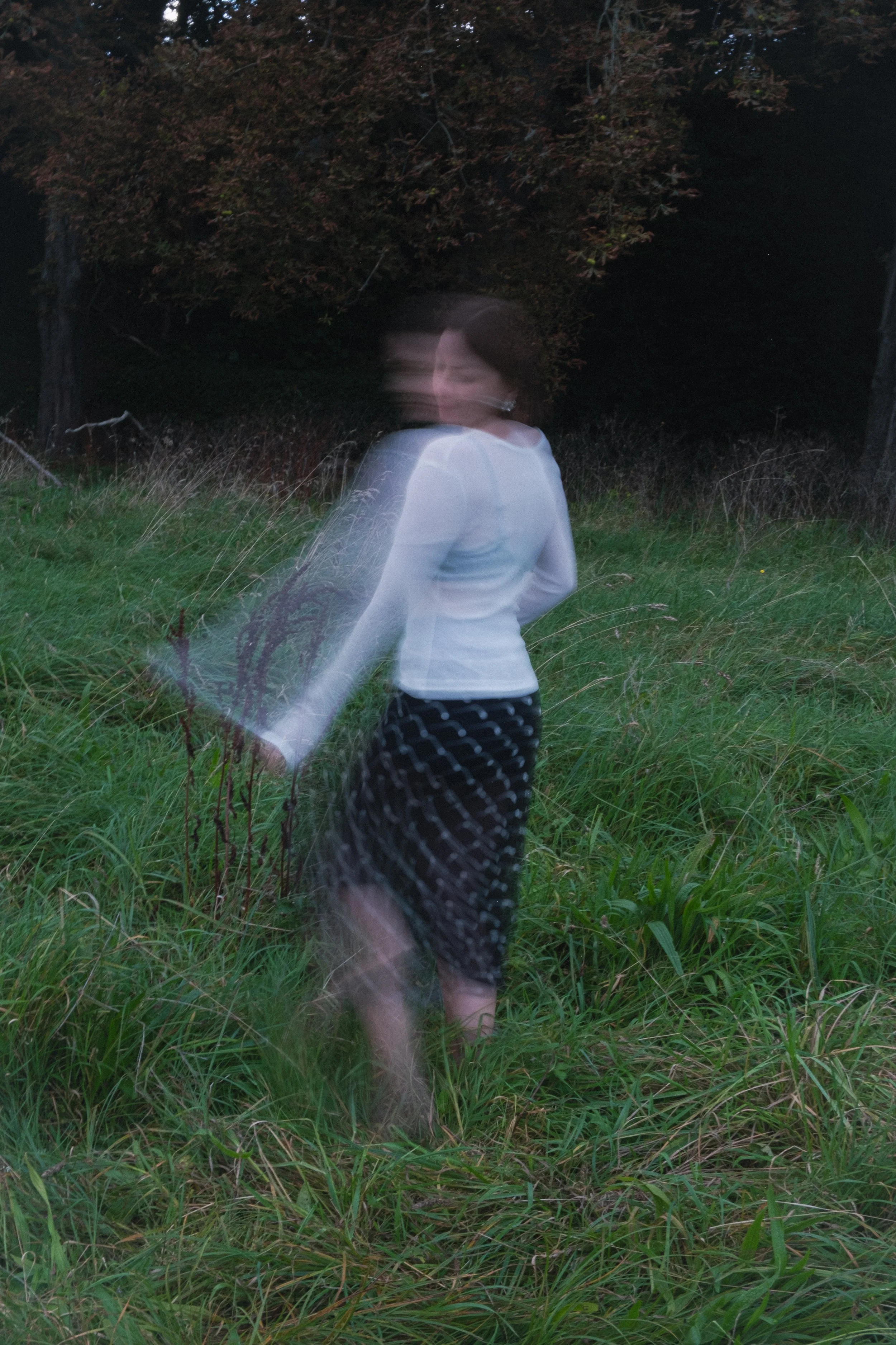 A woman in a white top and black skirt standing in tall grass in a forested area during dusk or dawn. The photo has motion blur, making her appear ghostly or in motion.