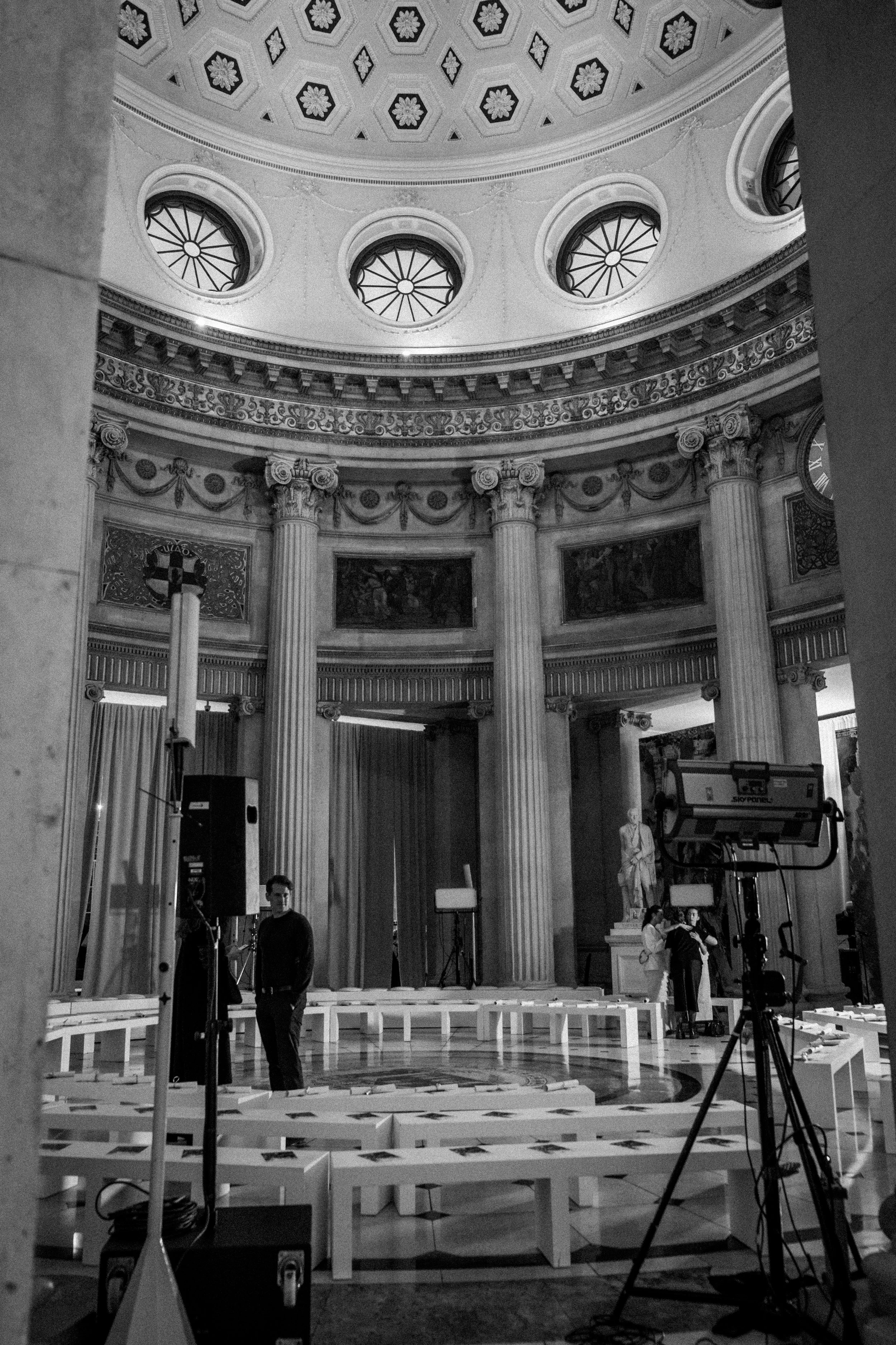 Black and white photo of an ornate, classical interior with tall Corinthian columns, a decorated dome ceiling with circular windows, and a stage with stage lighting and equipment. Several people are present, possibly preparing for an event.