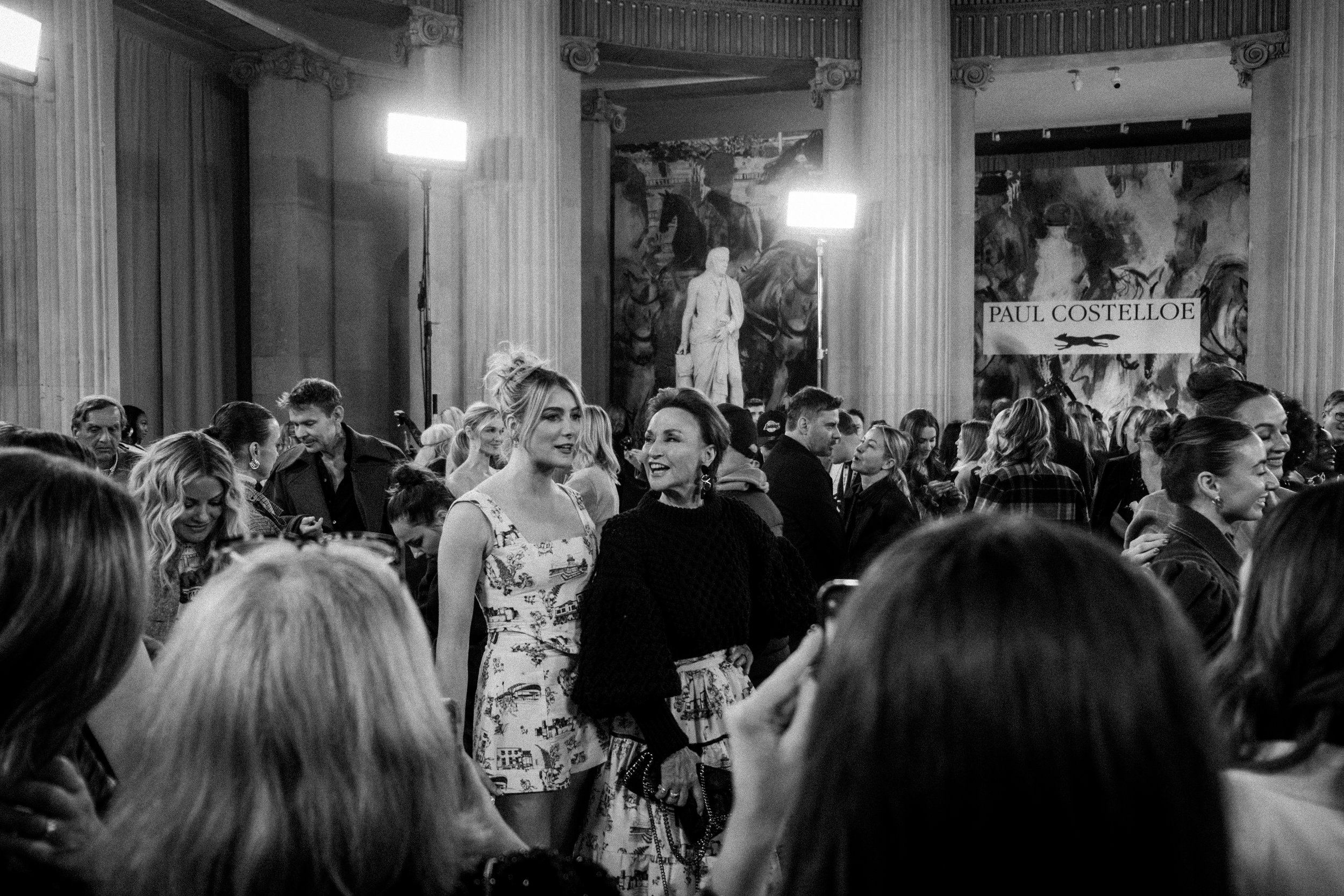 Black and white photo of a crowded event inside a hall with large columns and artwork, featuring two women in conversation in the center.
