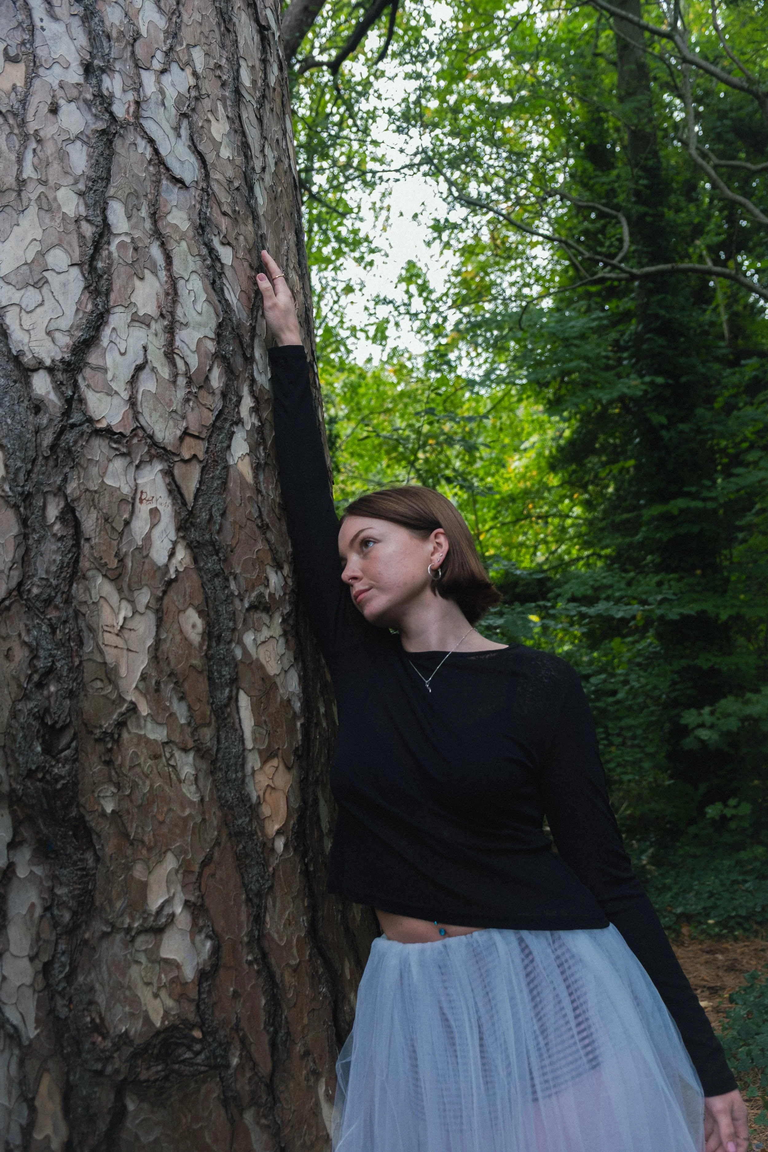 A woman wearing a black long-sleeve top and white tulle skirt standing next to a large tree with textured bark in a green forest.
