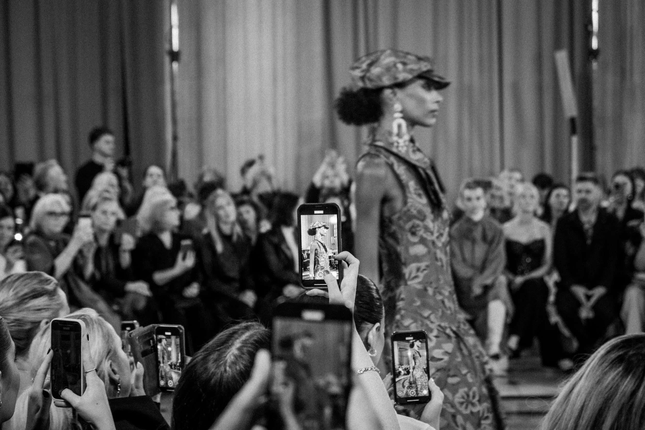 A black and white photo of a fashion show, with a model walking down the runway wearing a hat and patterned dress, while audience members in the background and foreground are taking pictures with their smartphones.