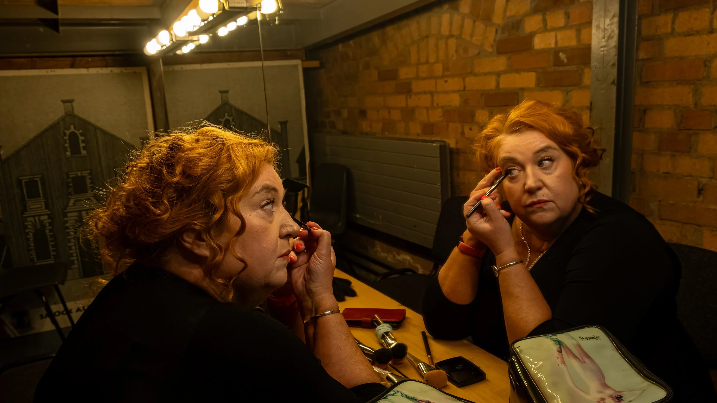 Two women with curly red hair sitting at a table, applying makeup while looking into a mirror.