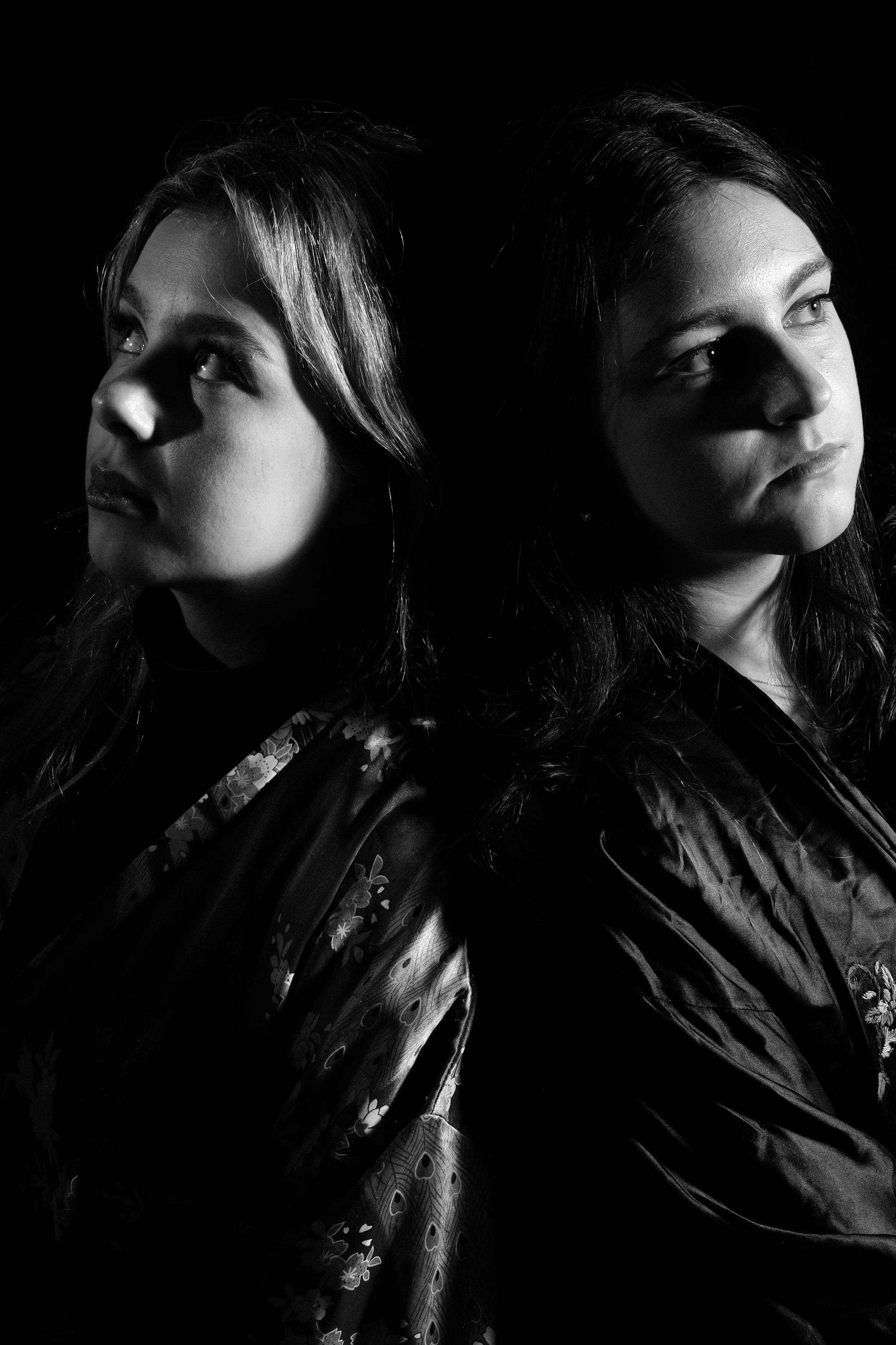Black and white photo of two women with shoulder-length hair, facing away from each other, looking in opposite directions, in dramatic lighting with shadows.