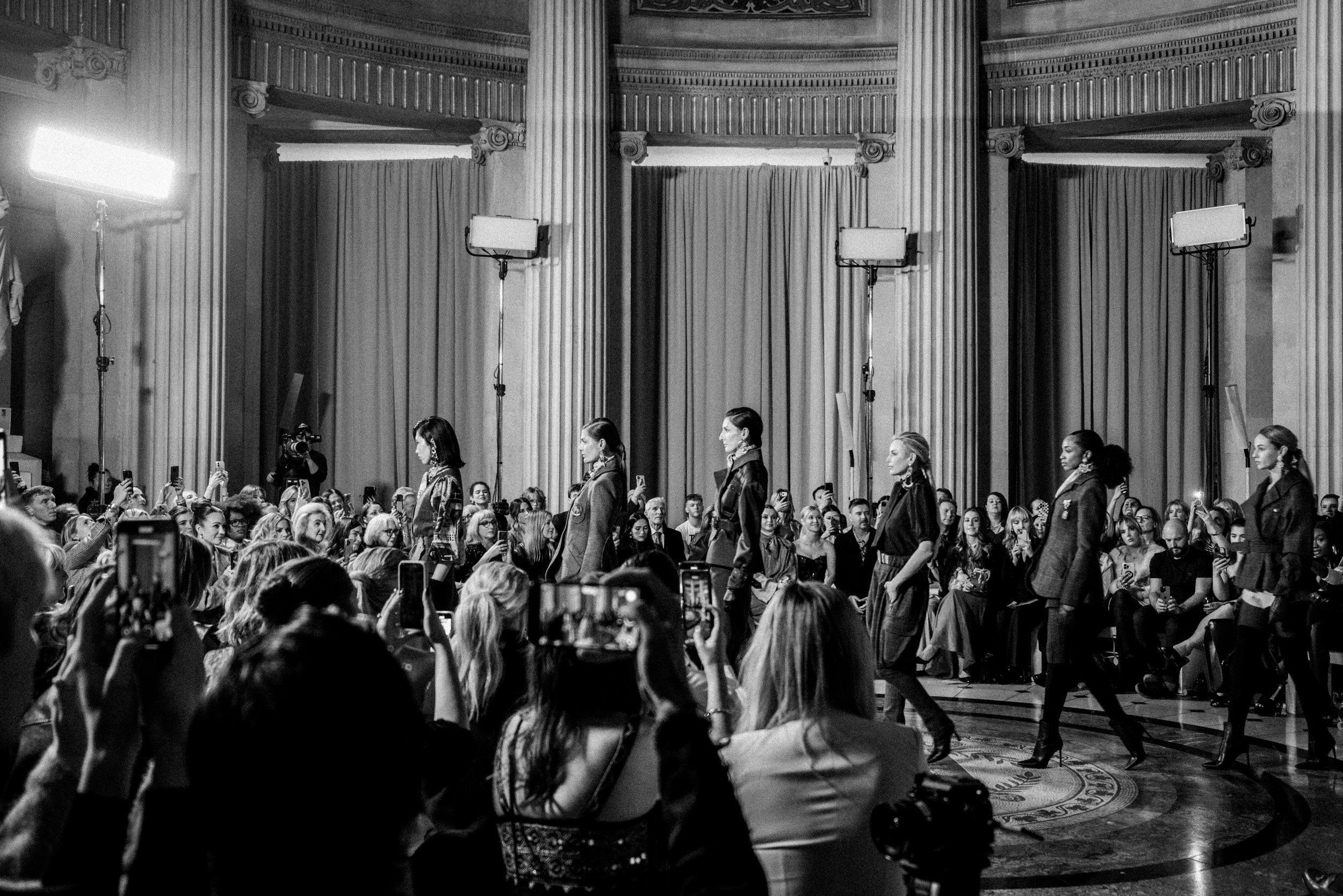 Black and white photo of a fashion runway show in a grand hall with tall columns and curtains. Models walk on the stage, while audience members and photographers are capturing the event.