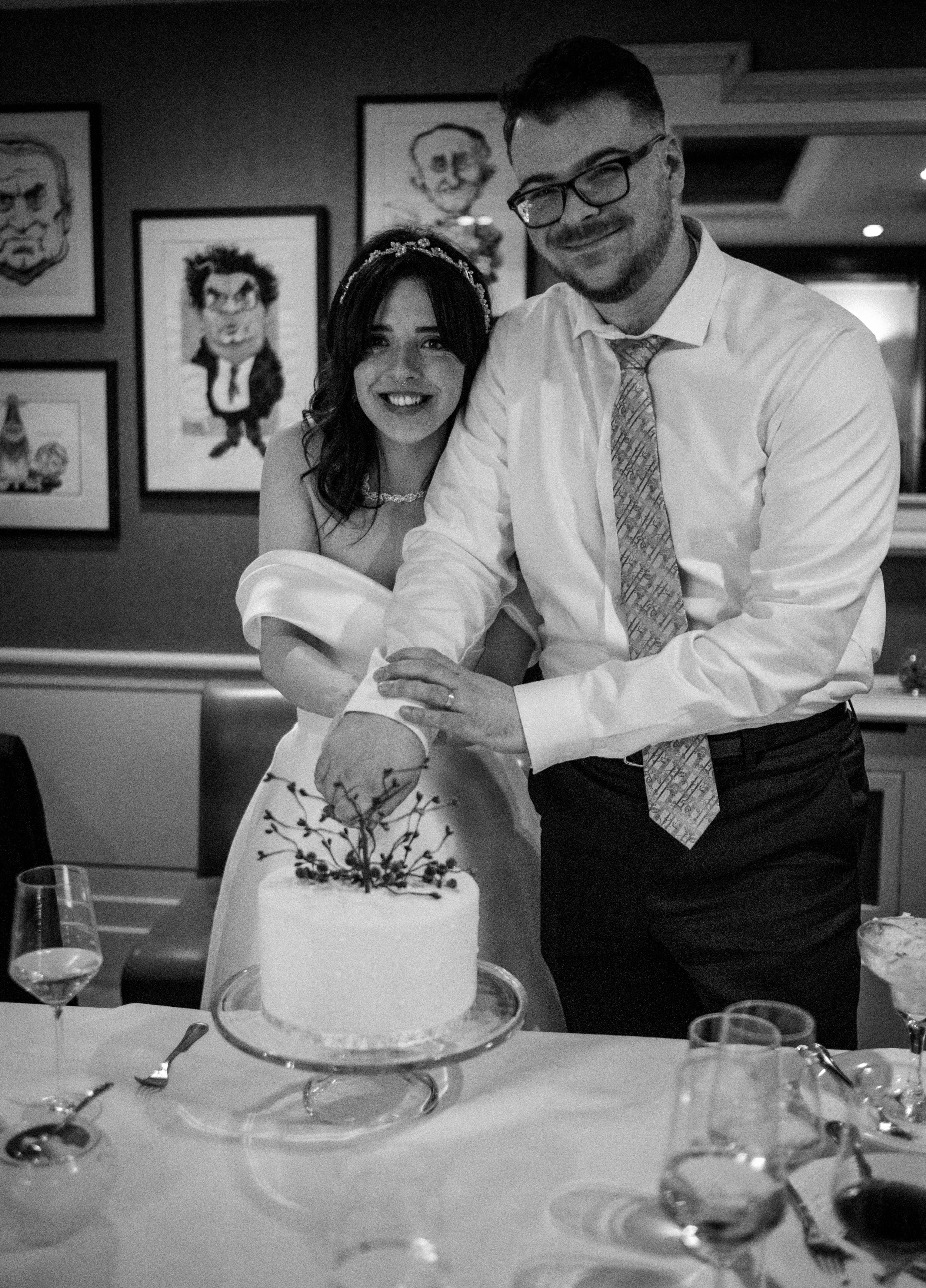 A bride and groom cutting a wedding cake together at a reception. The bride has dark hair, wears a tiara, and a white wedding dress. The groom has glasses, a beard, and is dressed in a white shirt and patterned tie. There are glasses and utensils on 
