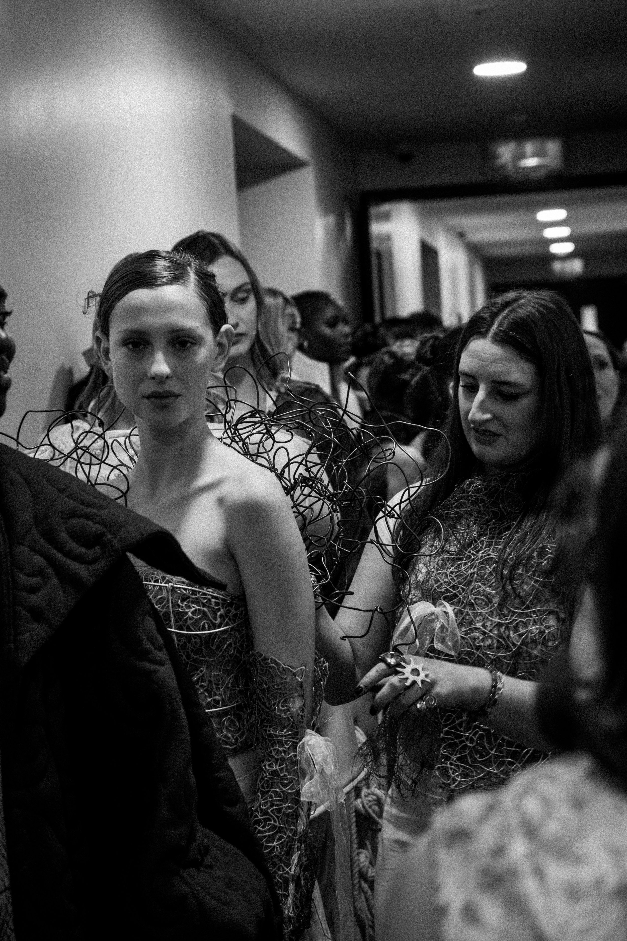 Group of women standing in a dimly lit hallway, with one woman in the foreground wearing a complex, wire-like outfit, and others in the background, some looking at her and some looking elsewhere.