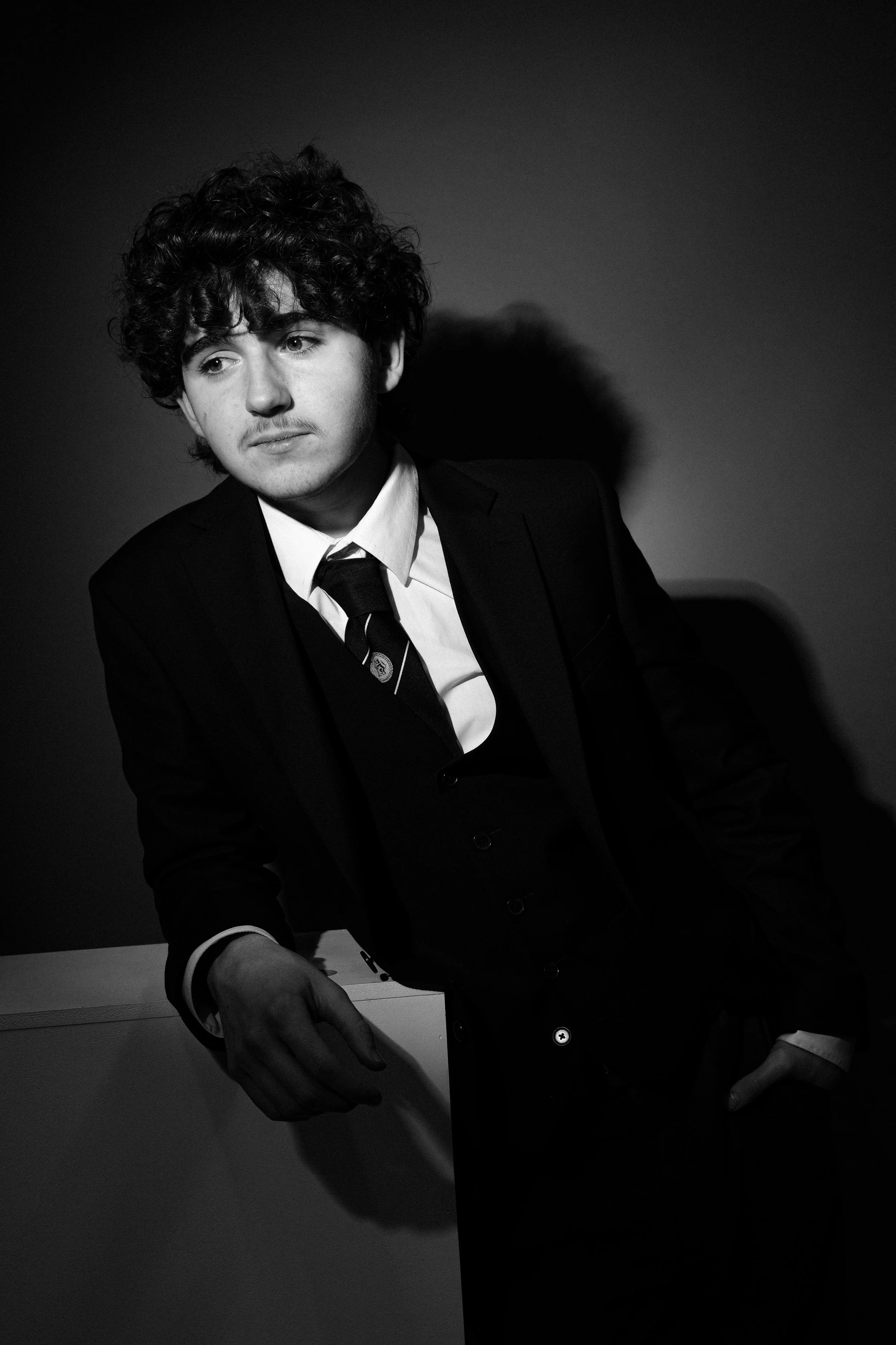 Black and white portrait of a young man with curly hair, wearing a suit and tie, leaning against a wall.