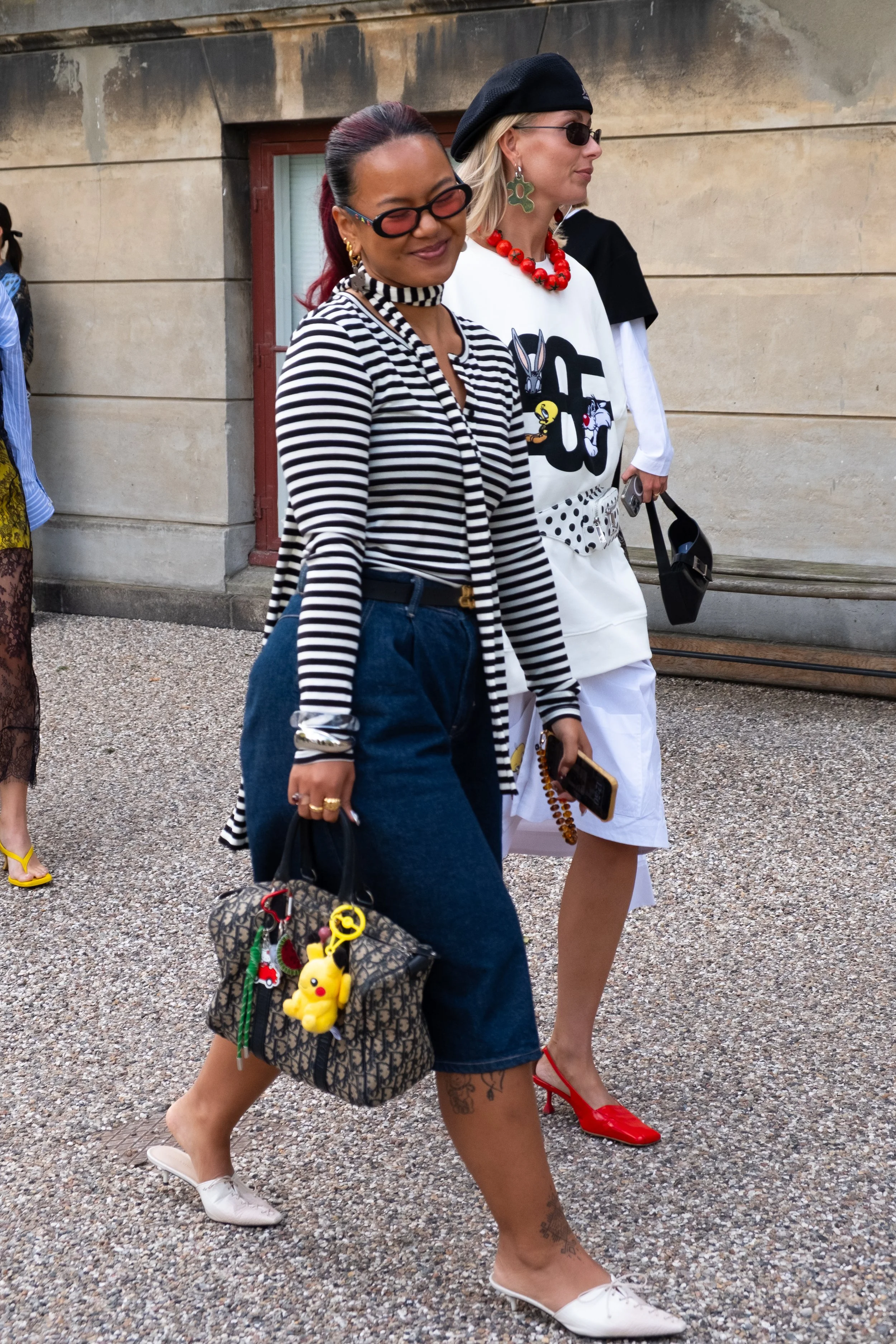 Two women walking outdoors. The woman in the foreground has dark red hair, glasses, and is wearing a black and white striped top, blue denim pants, and white shoes. She is carrying a designer bag with a yellow Pikachu keychain. The woman in the backg
