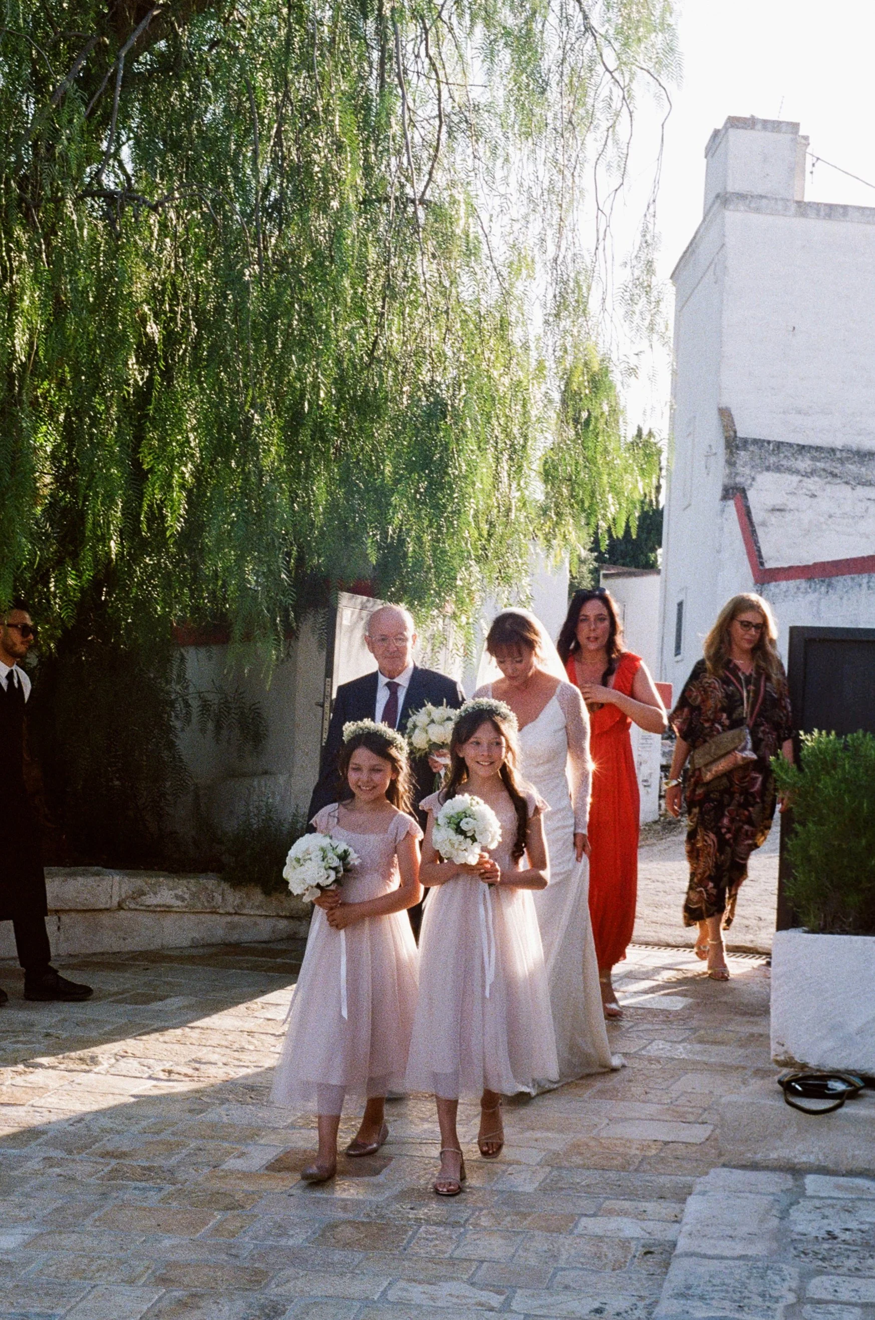 A wedding procession with a bride and two young flower girls walking outdoors, with bridesmaids and guests following, surrounded by trees and white buildings.