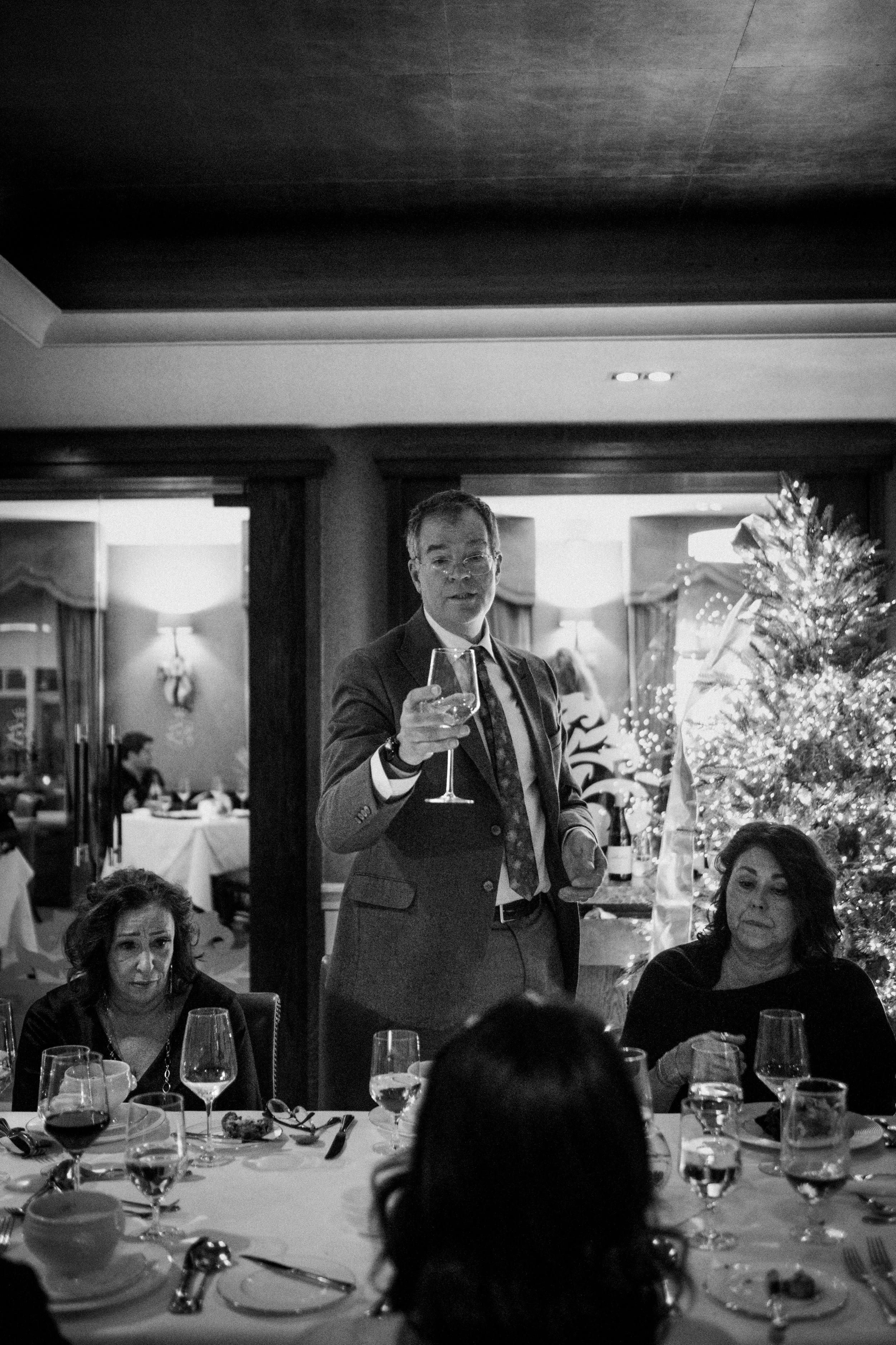 Black and white photo of a man in a suit standing at a dining table, making a toast with a wine glass during a Christmas gathering, with women seated and a decorated Christmas tree in the background.