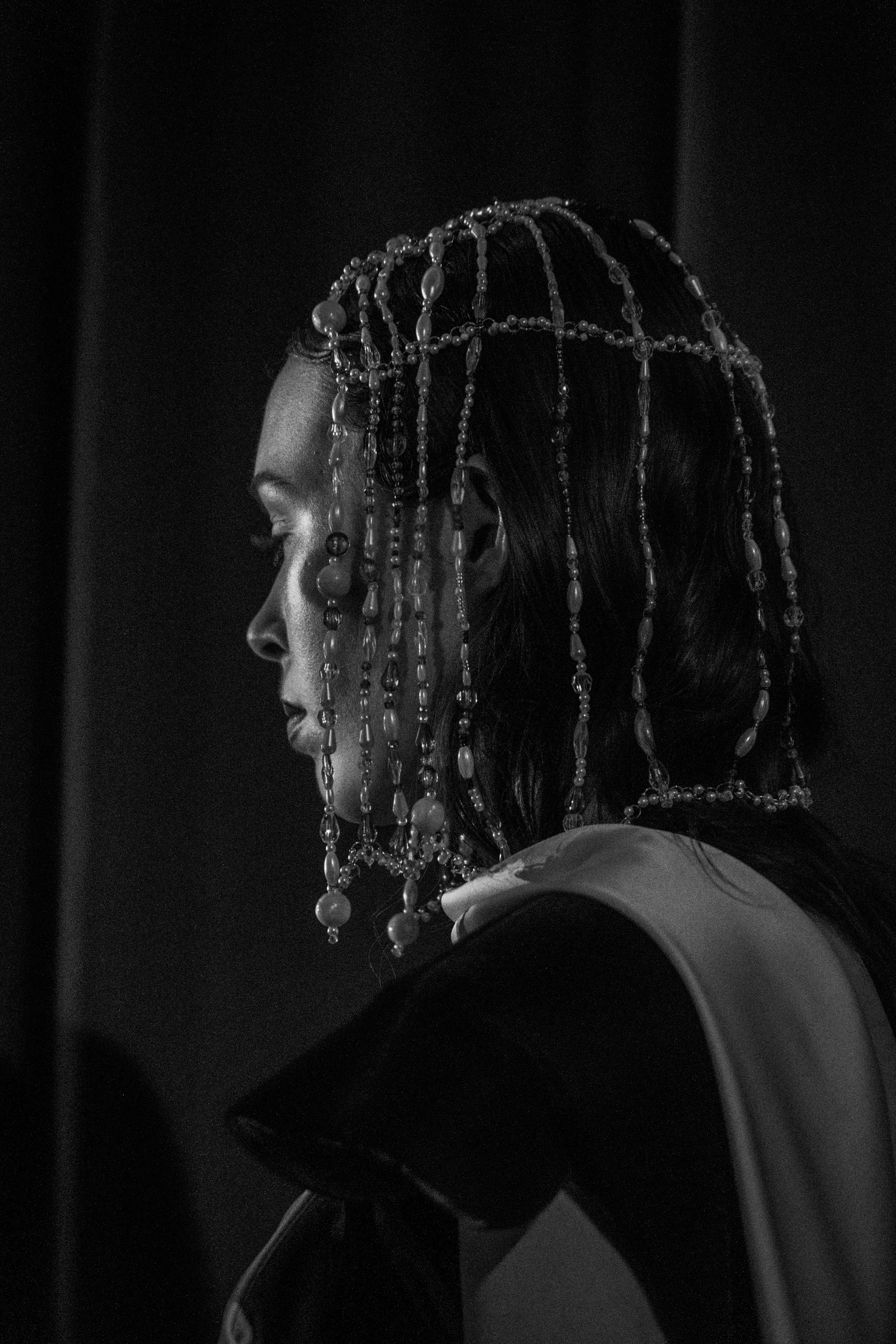 A woman in profile wearing an elaborate headdress made of beads and pearls, captured in black and white.