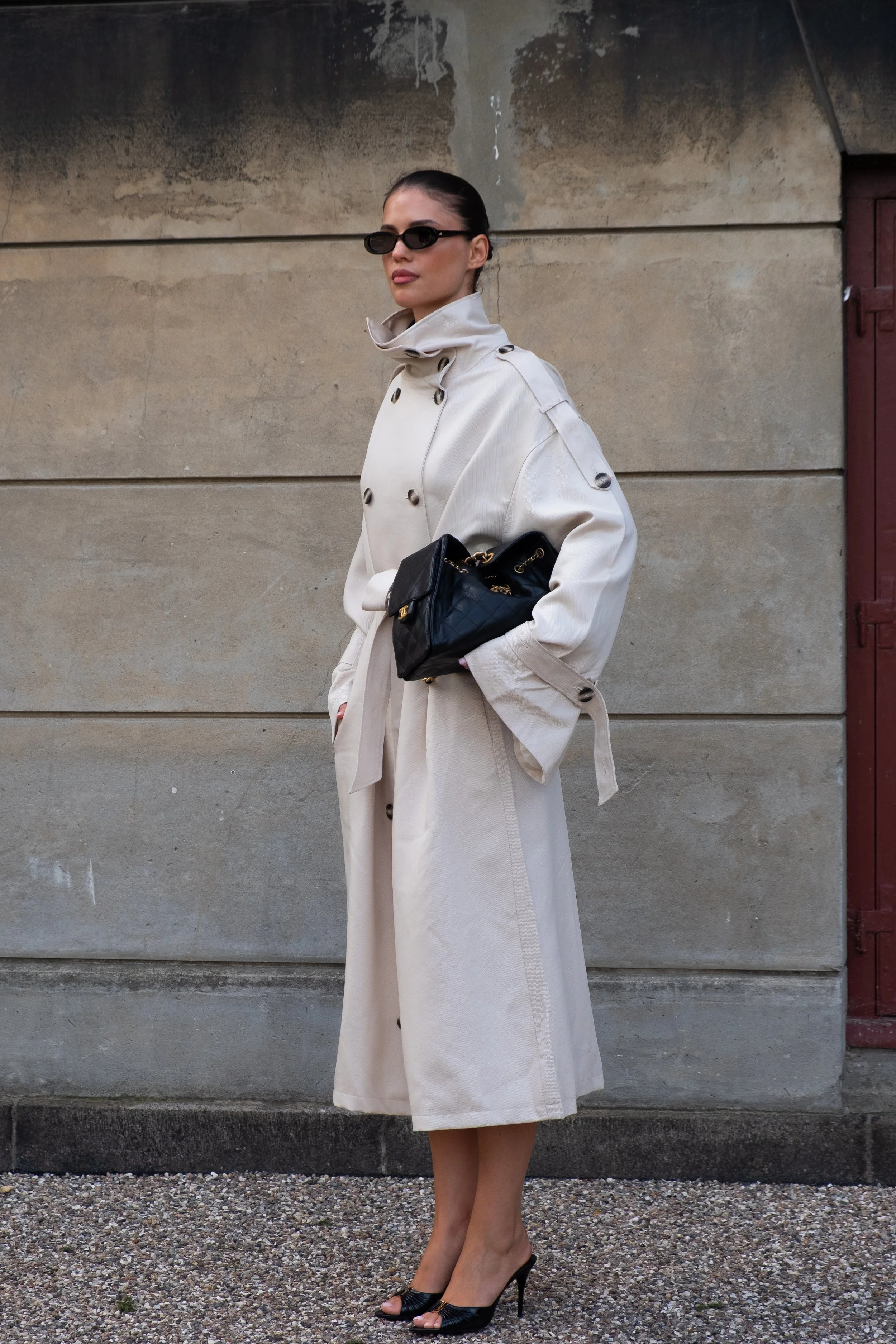 A woman wearing a beige trench coat, black sunglasses, and black high-heeled shoes, holding a black handbag, standing against a textured stone wall.