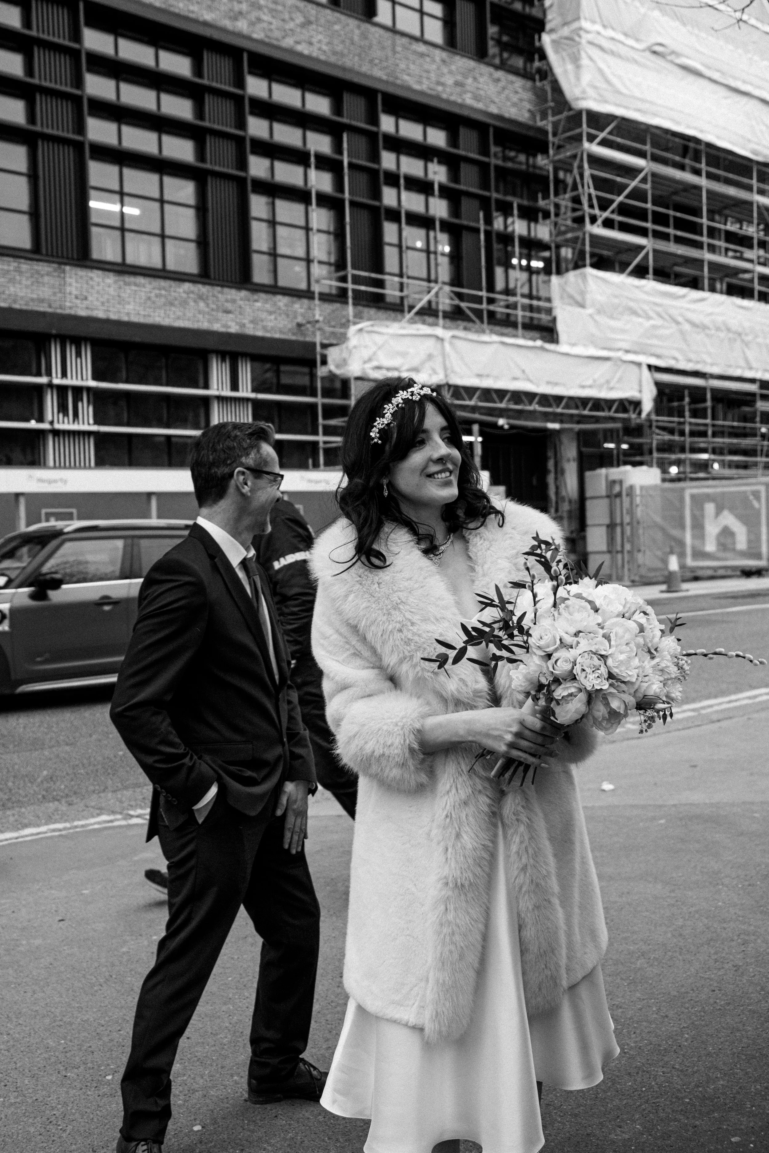 A woman in a wedding dress and fur coat holding a bouquet of flowers, smiling, standing on a city street. A man in a suit stands behind her, talking to someone. Construction scaffolding and a car are visible in the background.