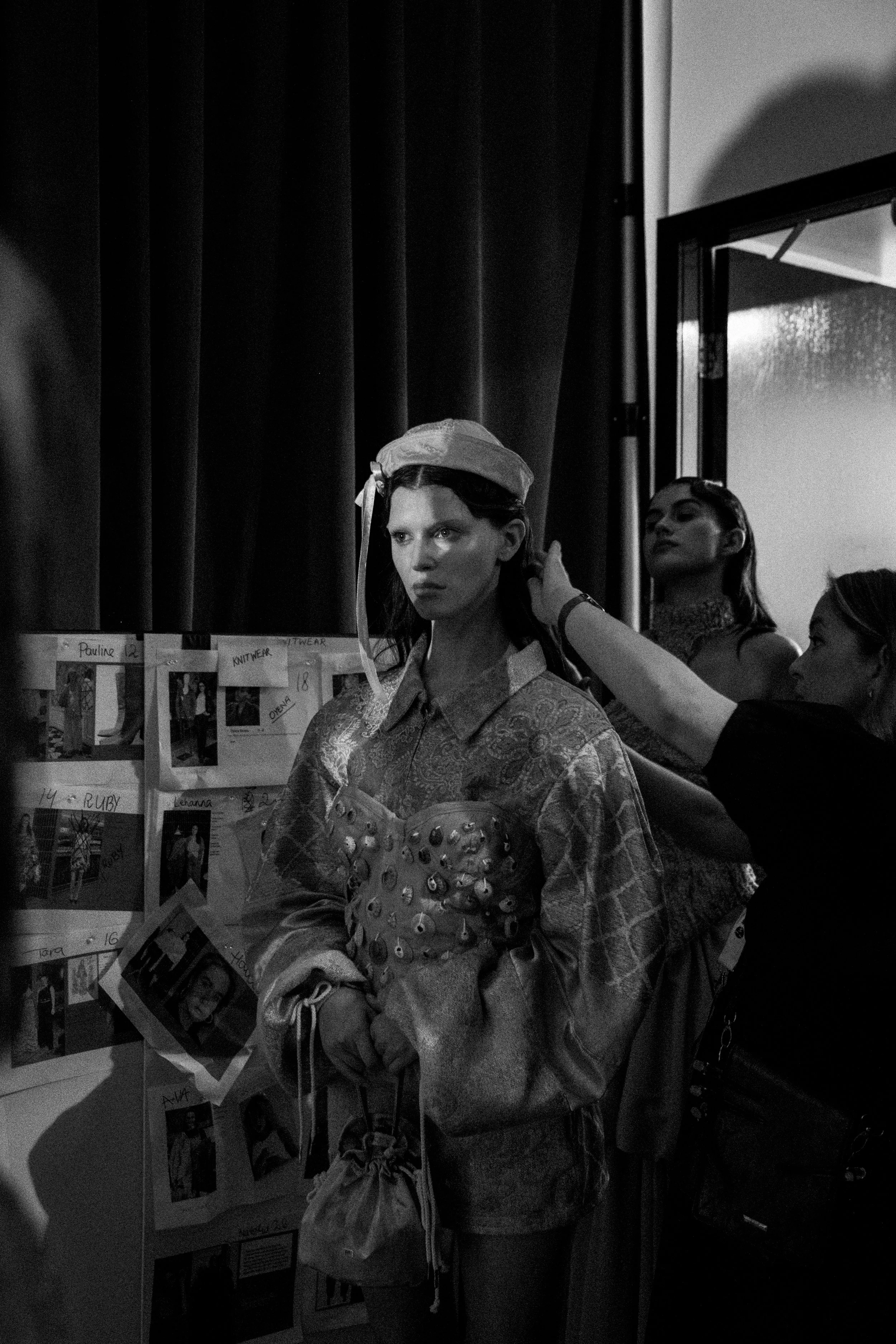 A woman in a patterned shirt is having her hair adjusted backstage at a fashion show, with two other women partially visible and a bulletin board with photographs and notes on the wall.