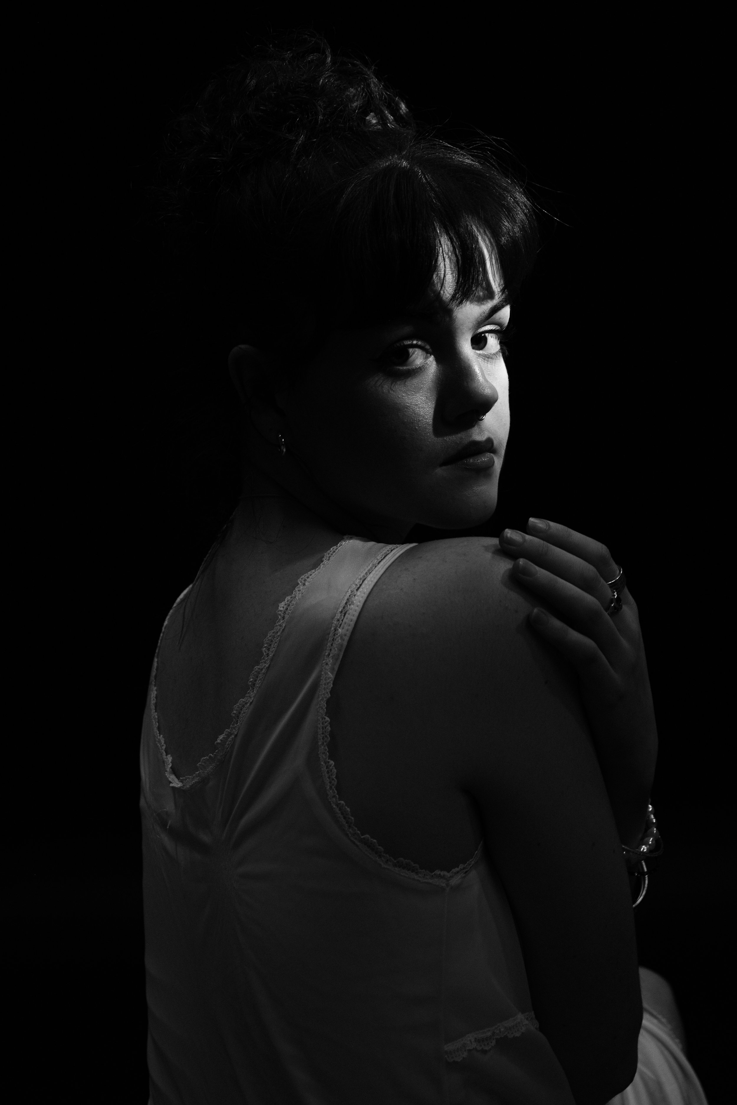 A black and white photo of a woman with dark hair styled in an updo, looking over her shoulder at the camera, with her hand resting on her shoulder, wearing a sleeveless top with lace trim.