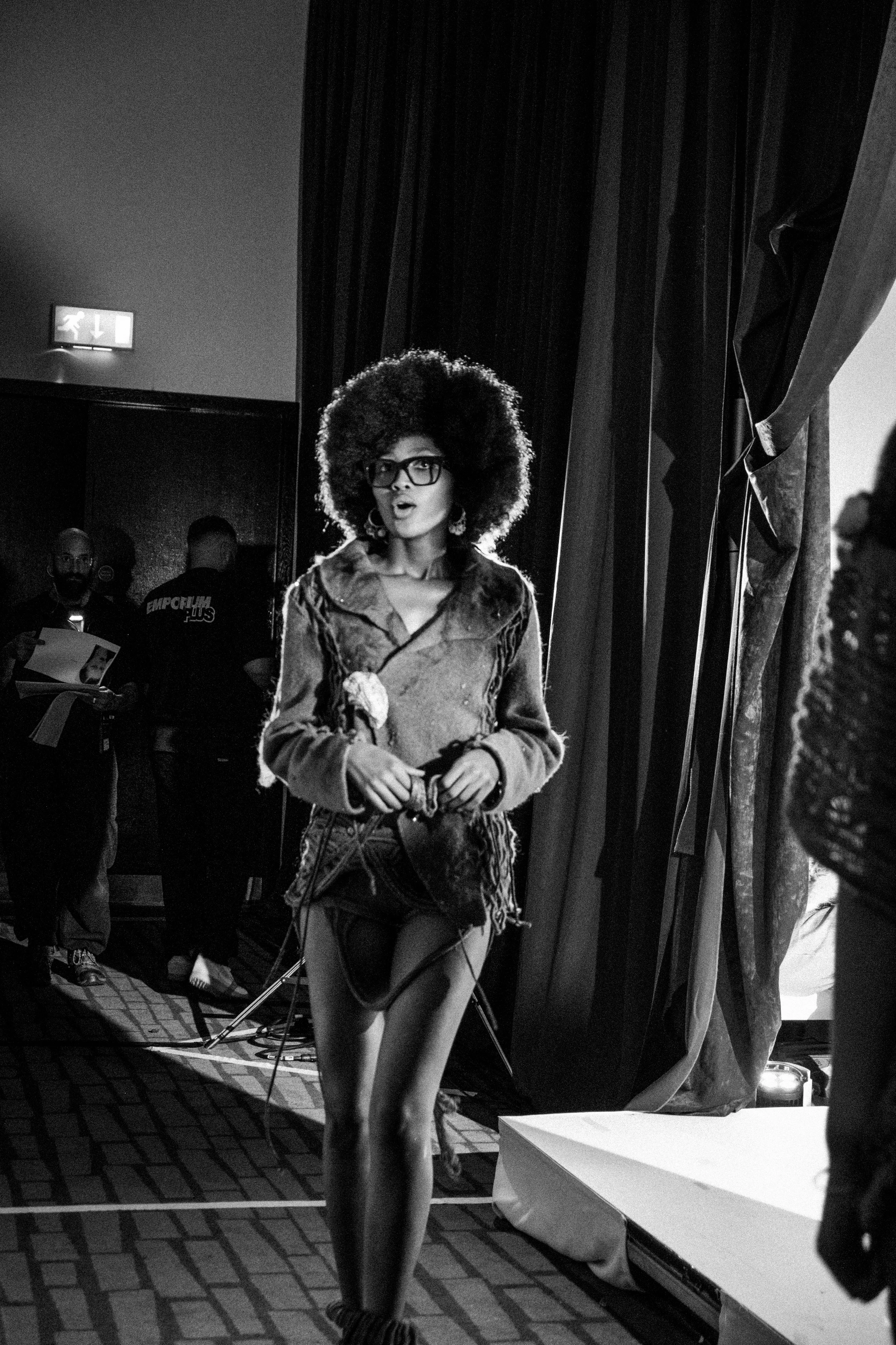 A woman with an afro hairstyle, glasses, and earrings, wearing a fur-lined coat and lace shorts, stands on a stage or backstage area, with a curtain and other people in the background.