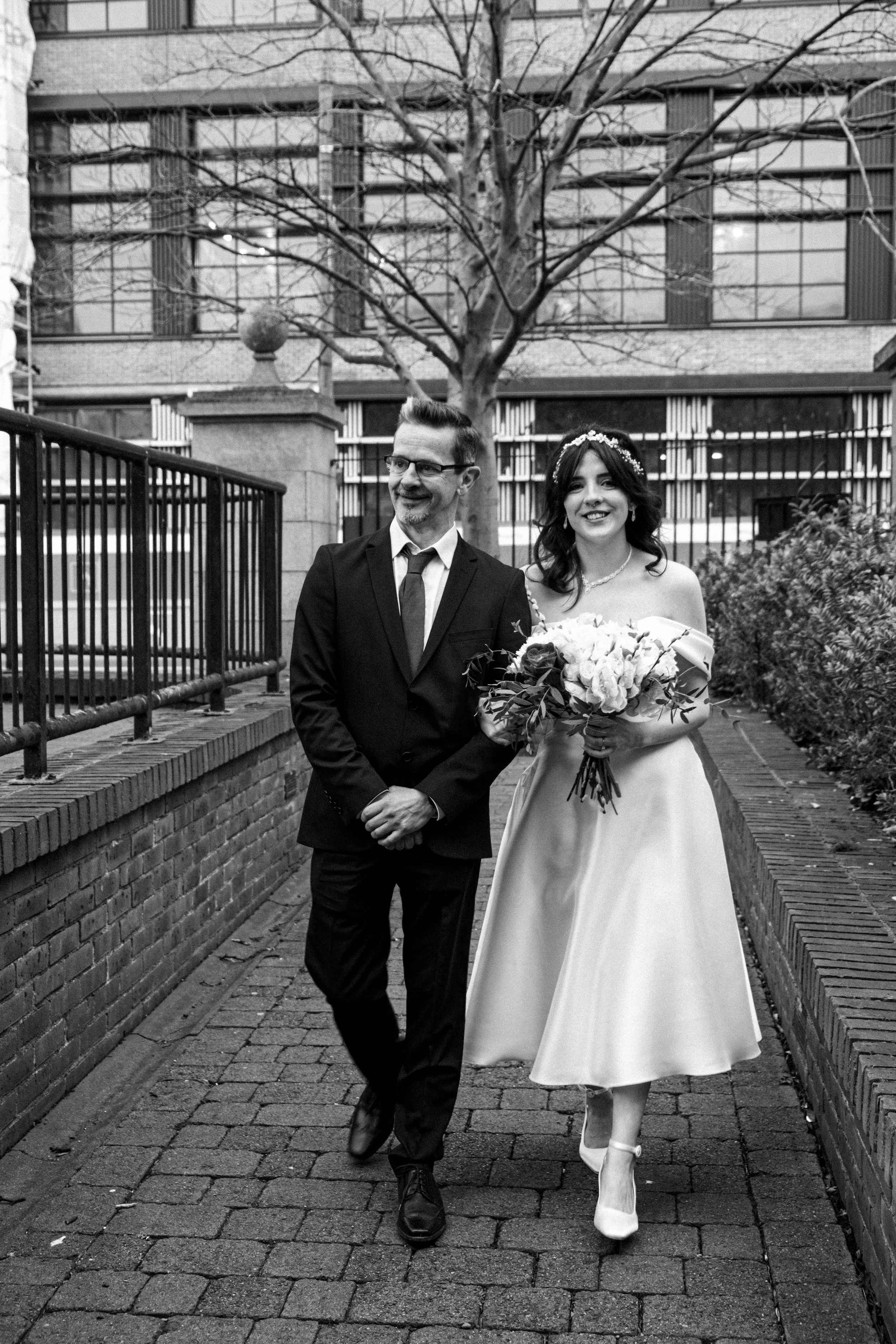 A black-and-white photo of a smiling bride holding a bouquet of flowers walking next to a man in a suit, possibly her father, outdoors on a brick pathway with bushes and a building in the background.
