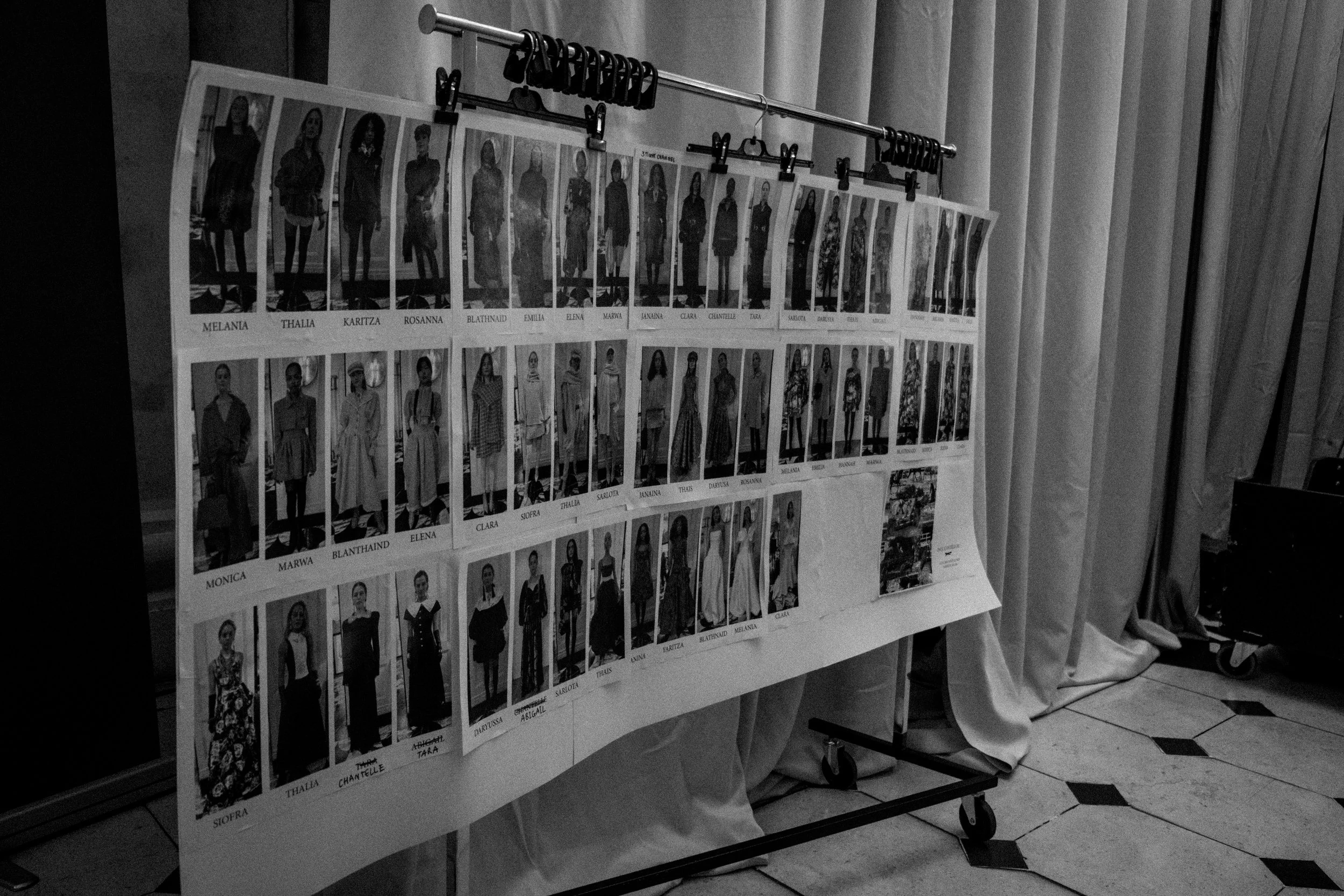 Display board with printed photos of individuals, each labeled with a name underneath, hanging on a stand with clips, set in a room with curtains and tiled floor.