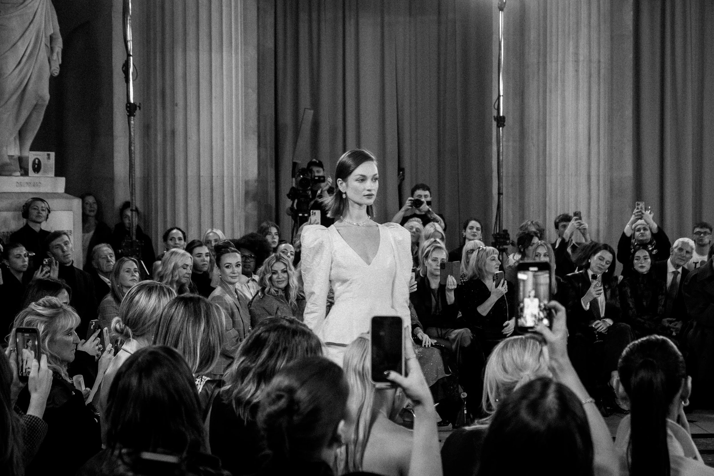A woman in a white dress walking on a runway at a fashion show, with an audience seated and standing around, many holding phones capturing photos.